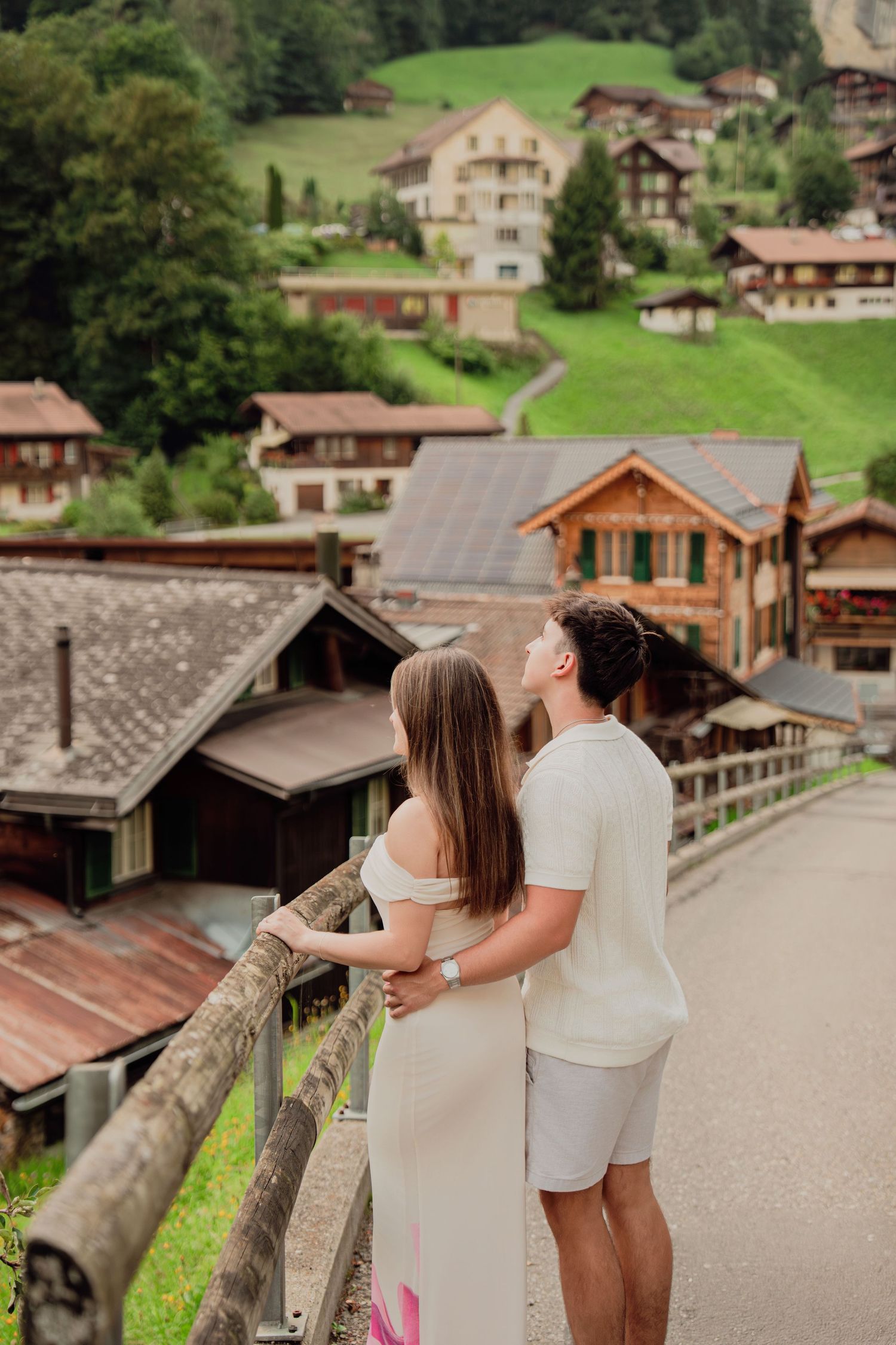 Romantic couple hugging on a wooden bridge overlooking traditional Swiss chalets on a green hillside.