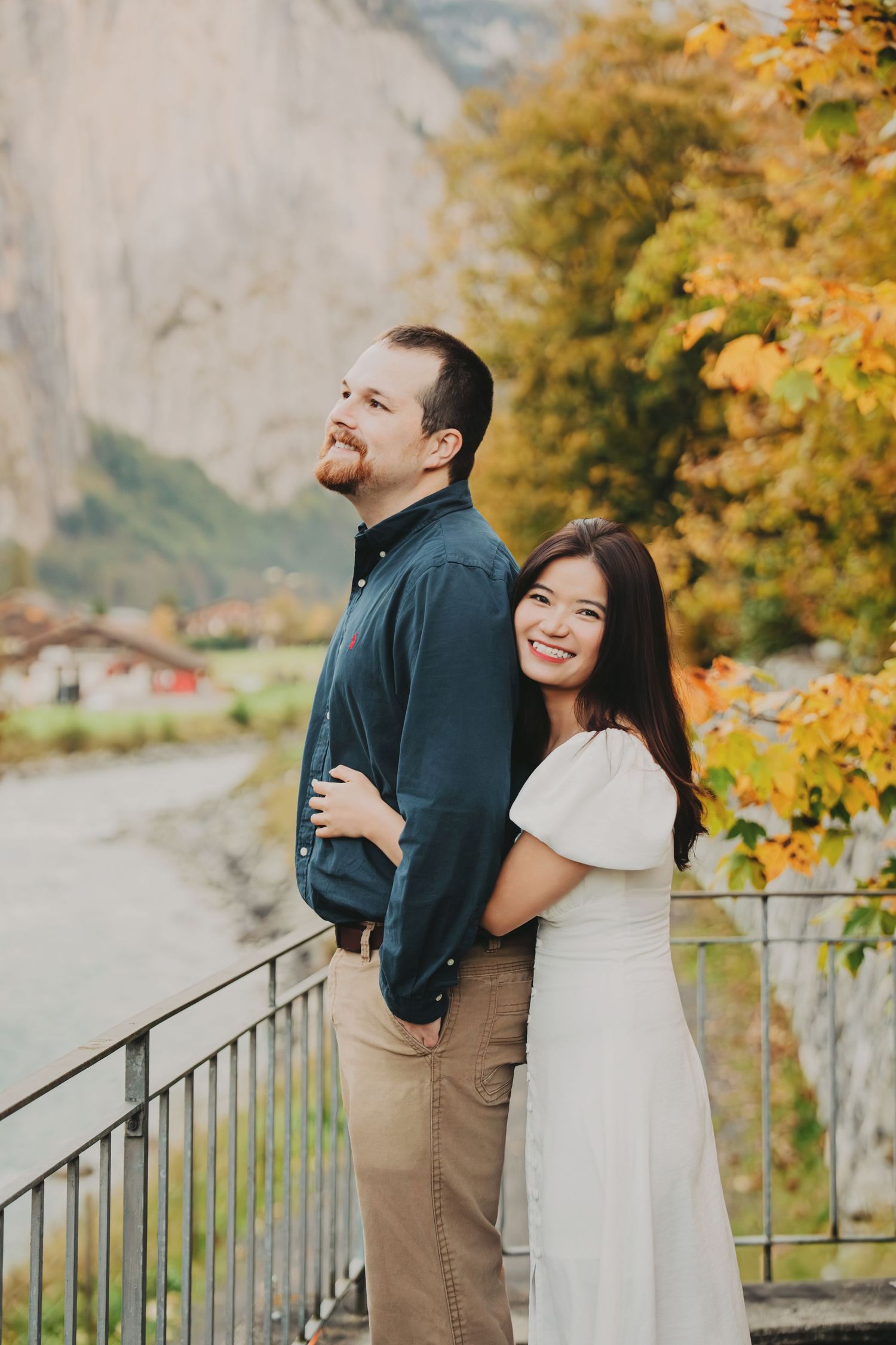 Woman hugging man from behind while smiling on a bridge with colorful fall trees.