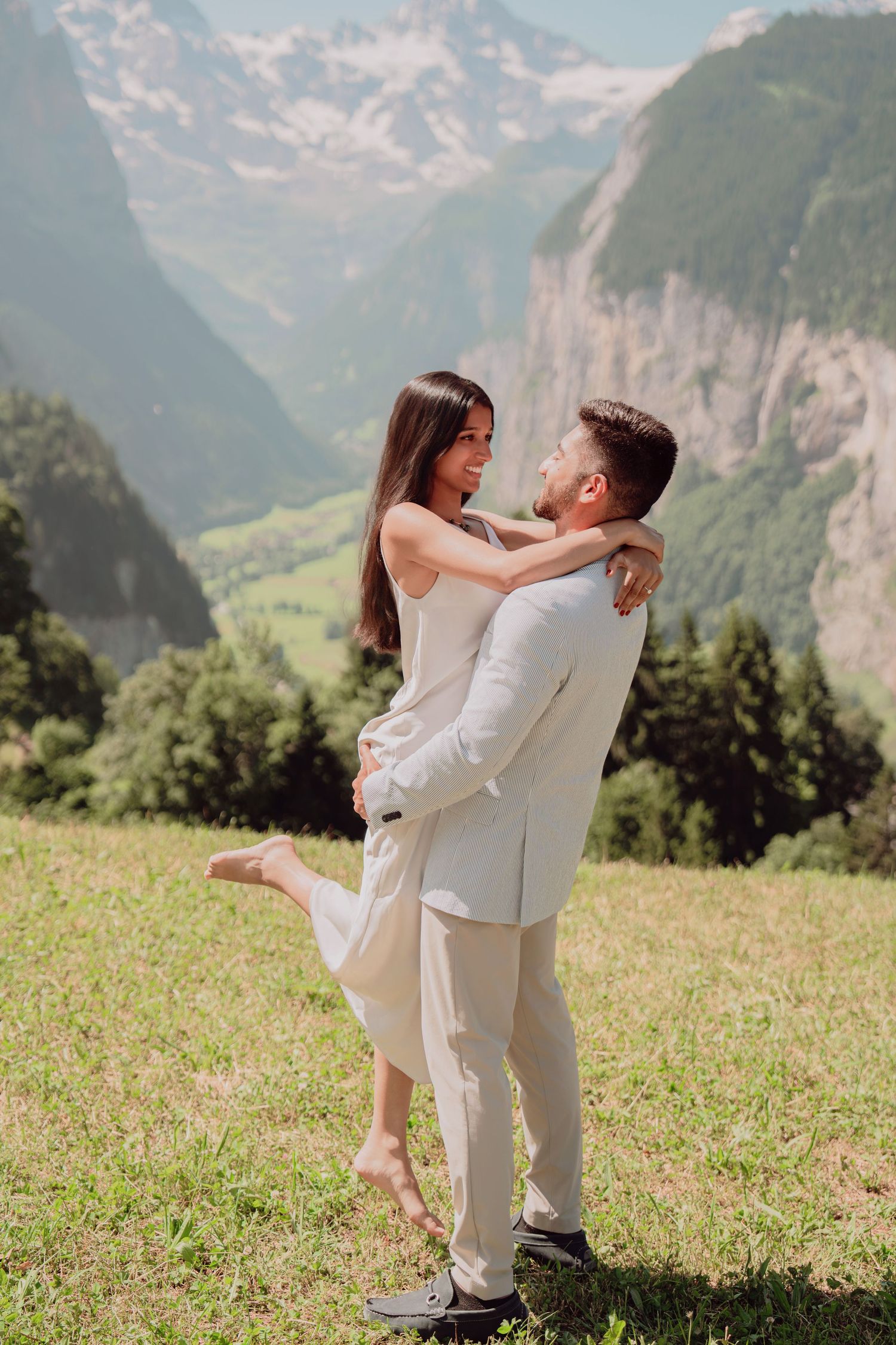 Couple embracing on green hillside with stunning Swiss Alpine valley and mountain backdrop.