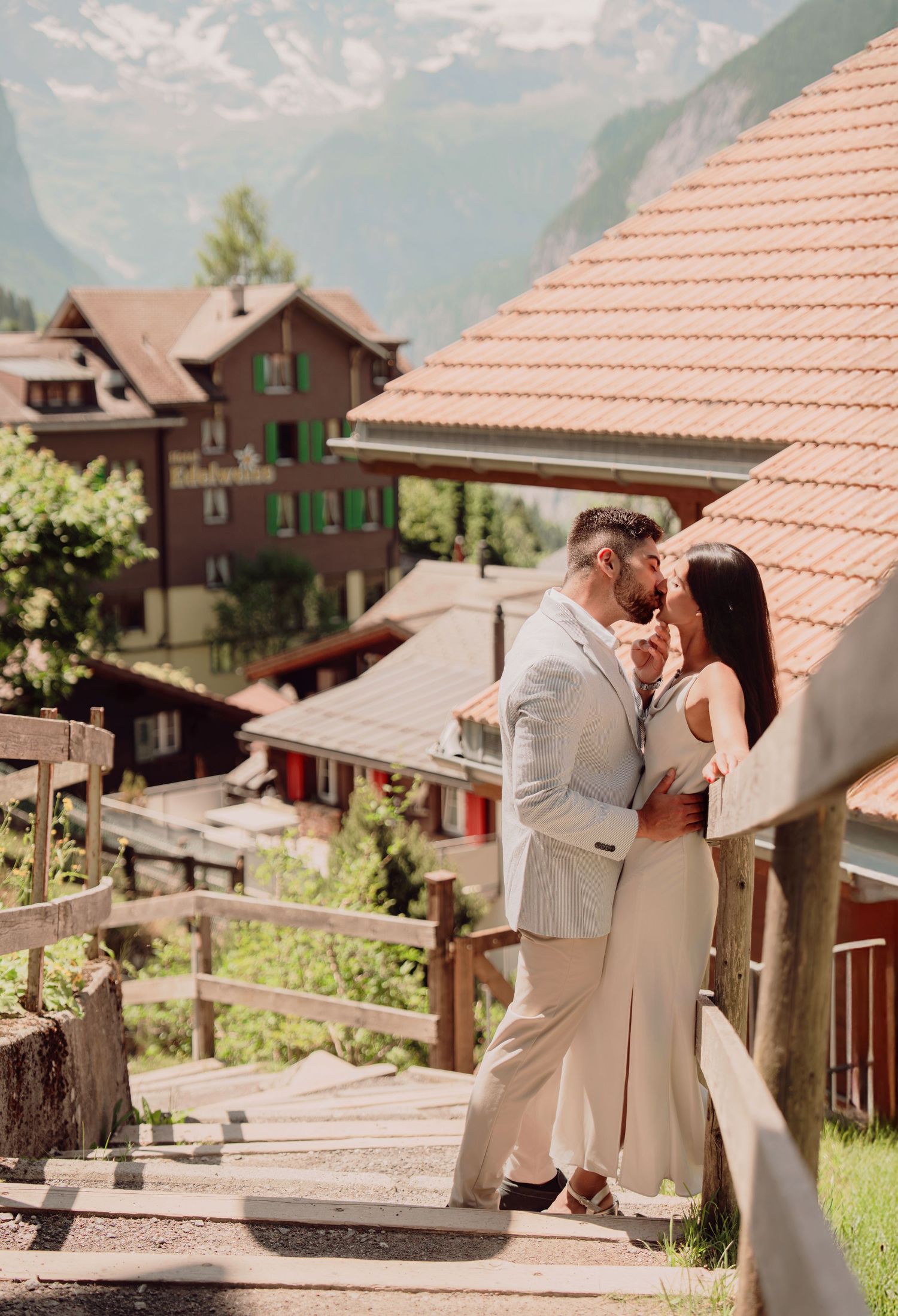 Couple sharing an intimate kiss on wooden stairs with charming Swiss chalet buildings and mountains in the background.