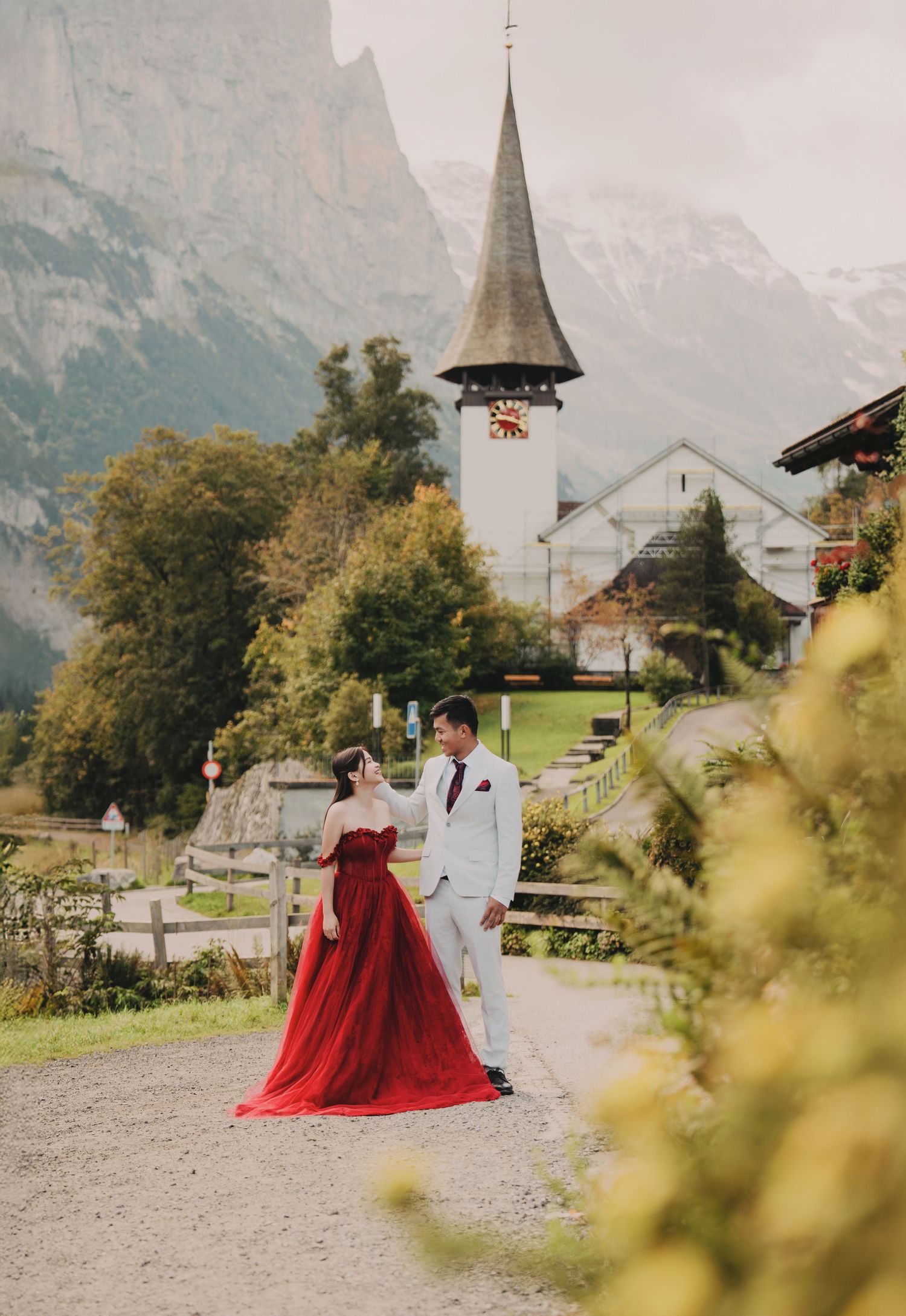 Couple in red dress and white suit walking near historic Swiss church with mountain backdrop.