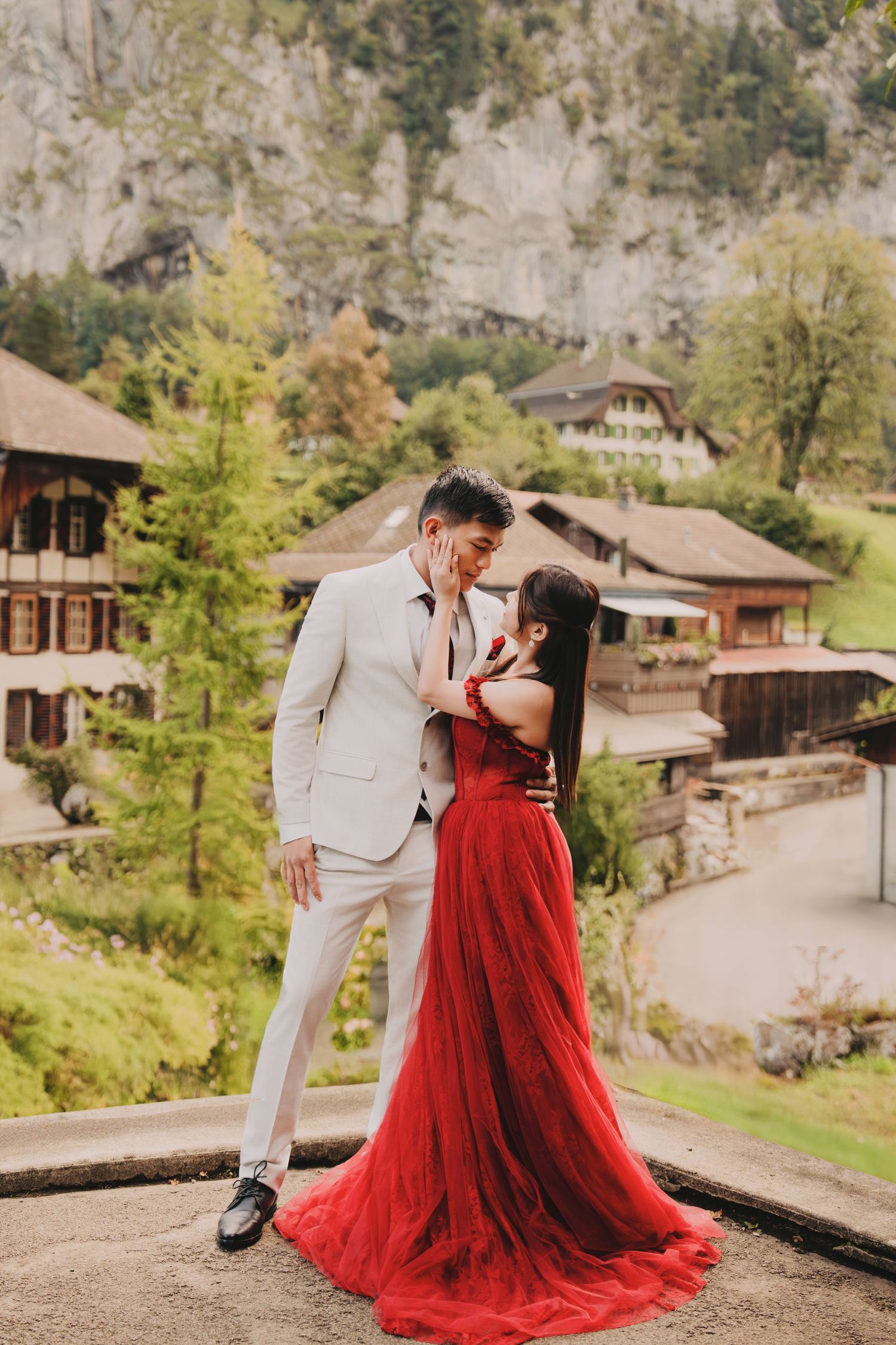 Bride in flowing red tulle gown and groom in white suit share intimate moment in Lauterbrunnen valley.