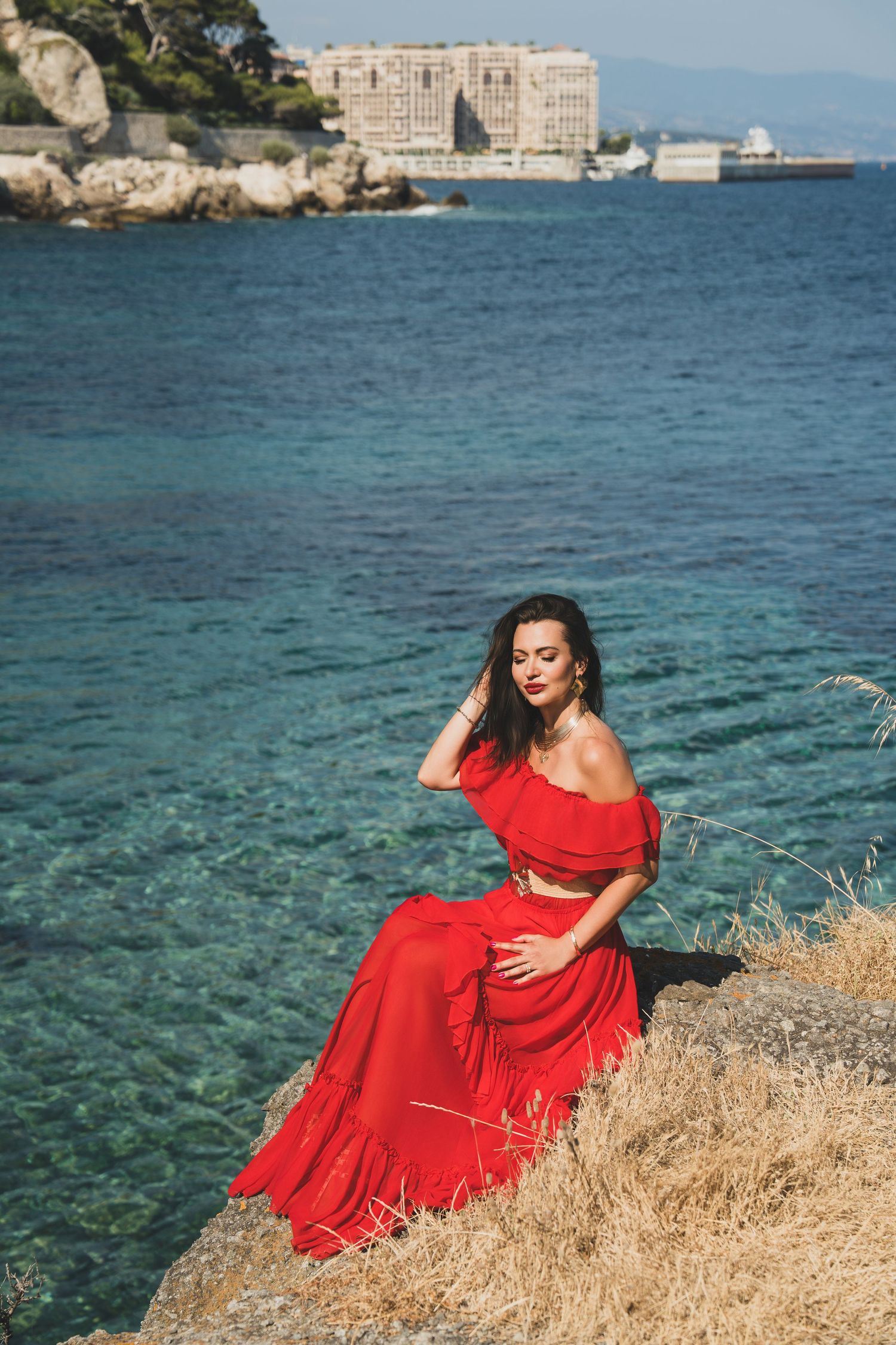 Woman in elegant red off-shoulder dress sitting on coastal rocks overlooking turquoise Mediterranean sea.