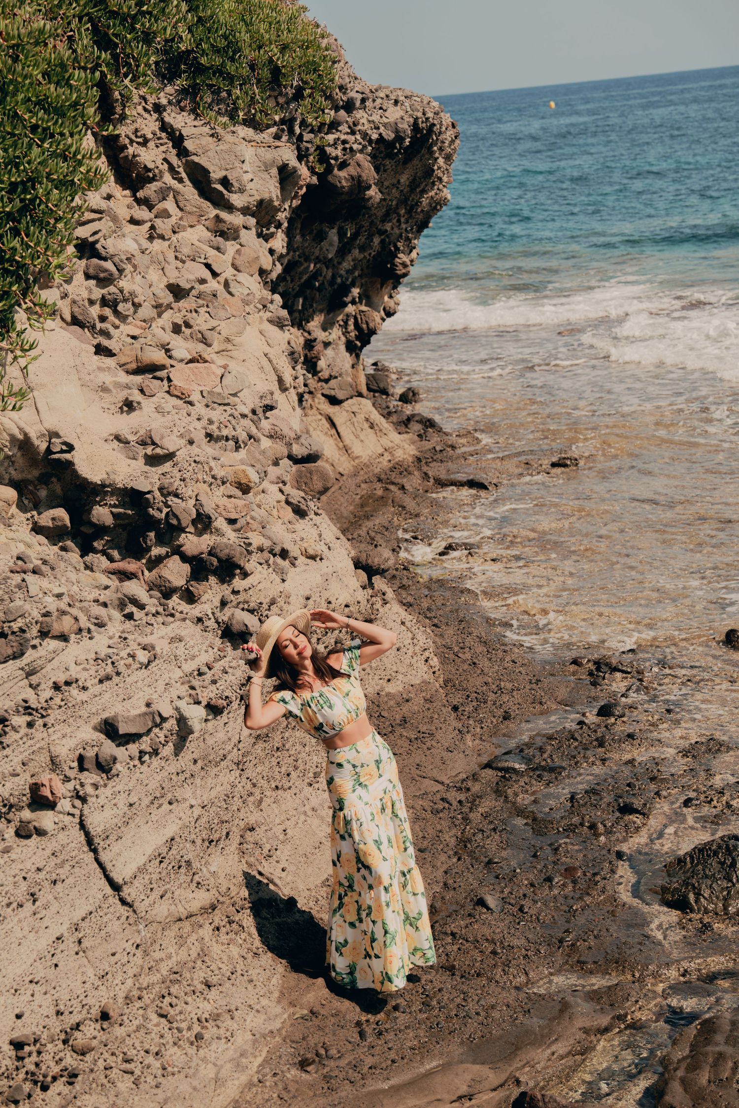 Woman in floral two-piece set posing against rocky seaside cliffs with ocean waves.