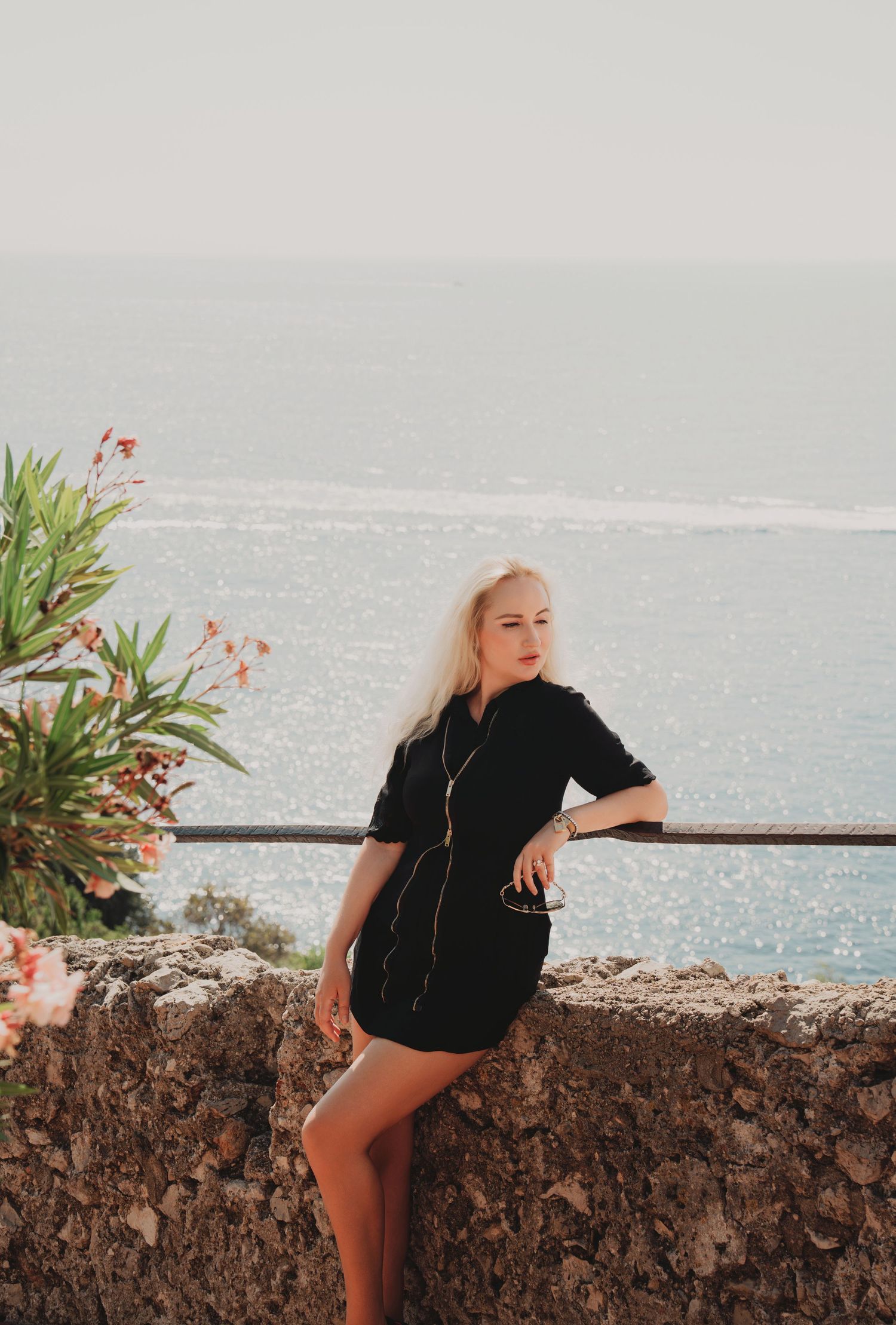 Blonde woman in black dress leaning against a stone wall with a stunning Mediterranean sea view behind her.