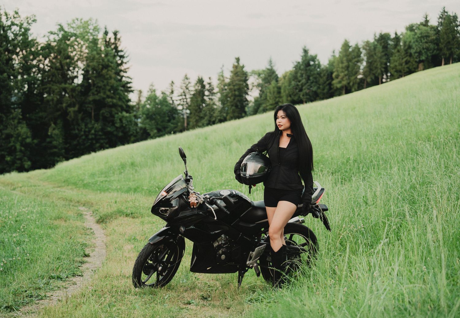 A woman in black outfit holds a helmet beside a black motorcycle on a green grassy hillside path.