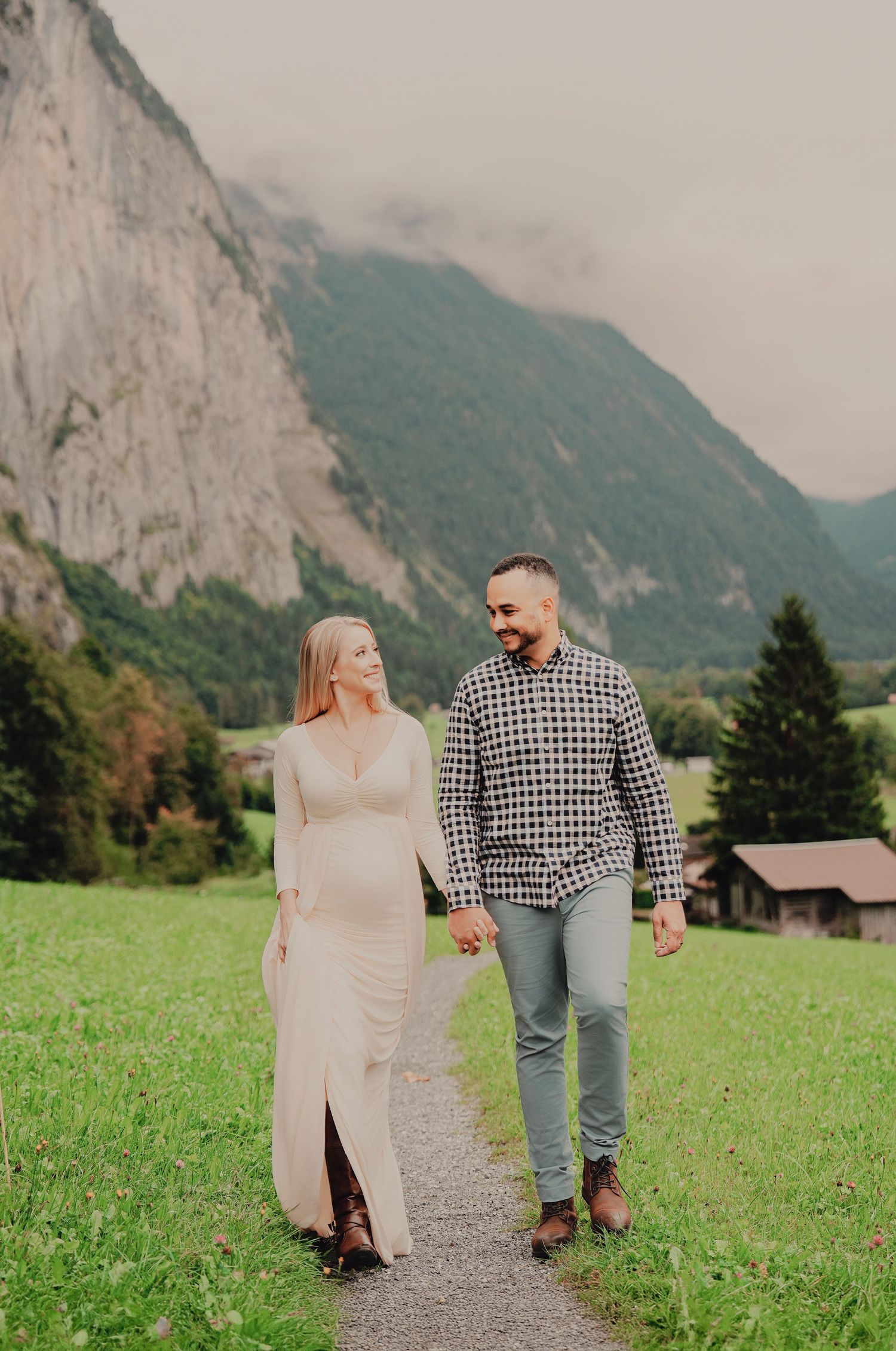 Smiling pregnant couple walks hand in hand on gravel path with dramatic Swiss mountain backdrop.