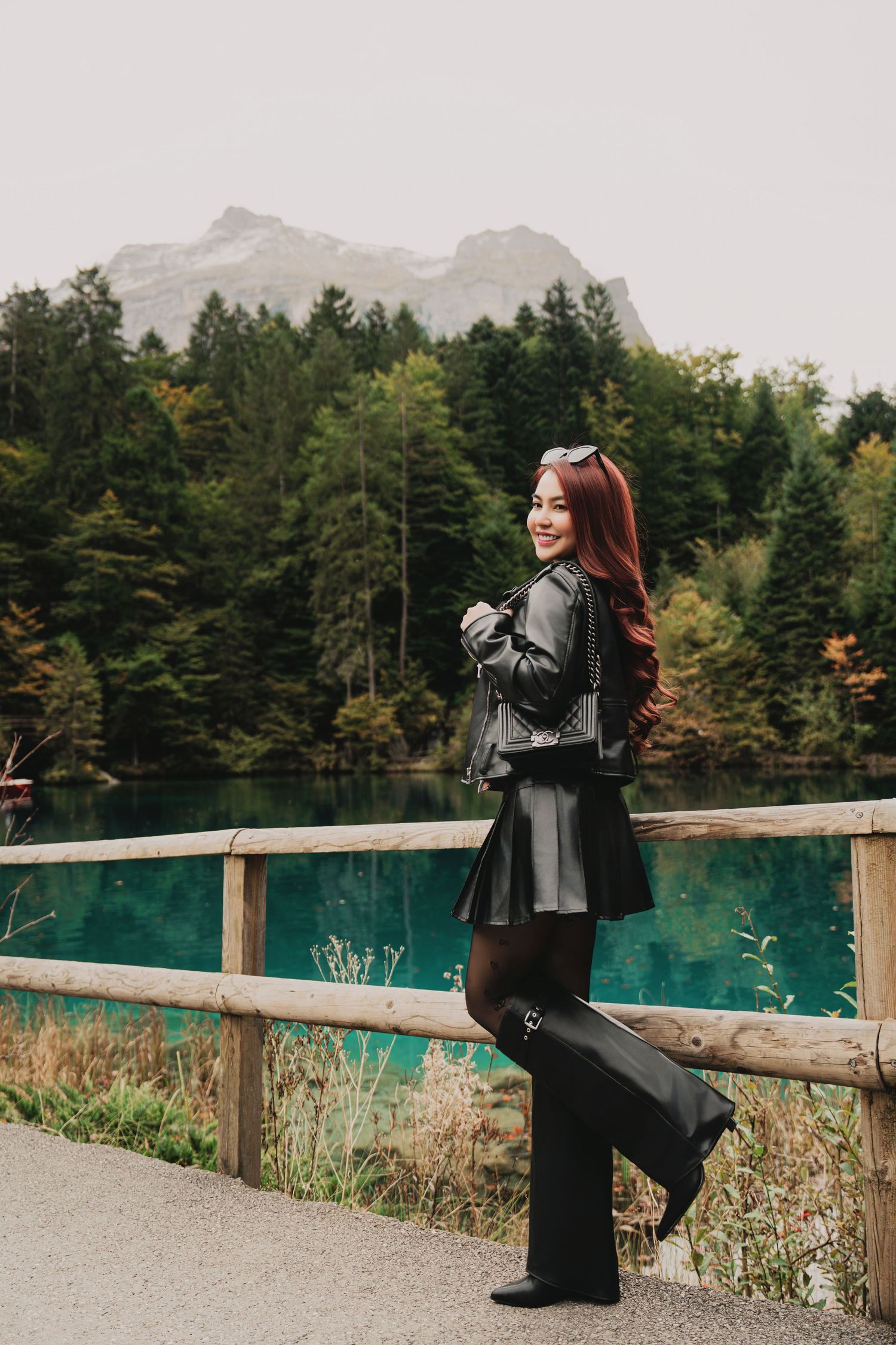 Cheerful red-haired woman in black leather outfit posing by turquoise lake with mountain scenery.