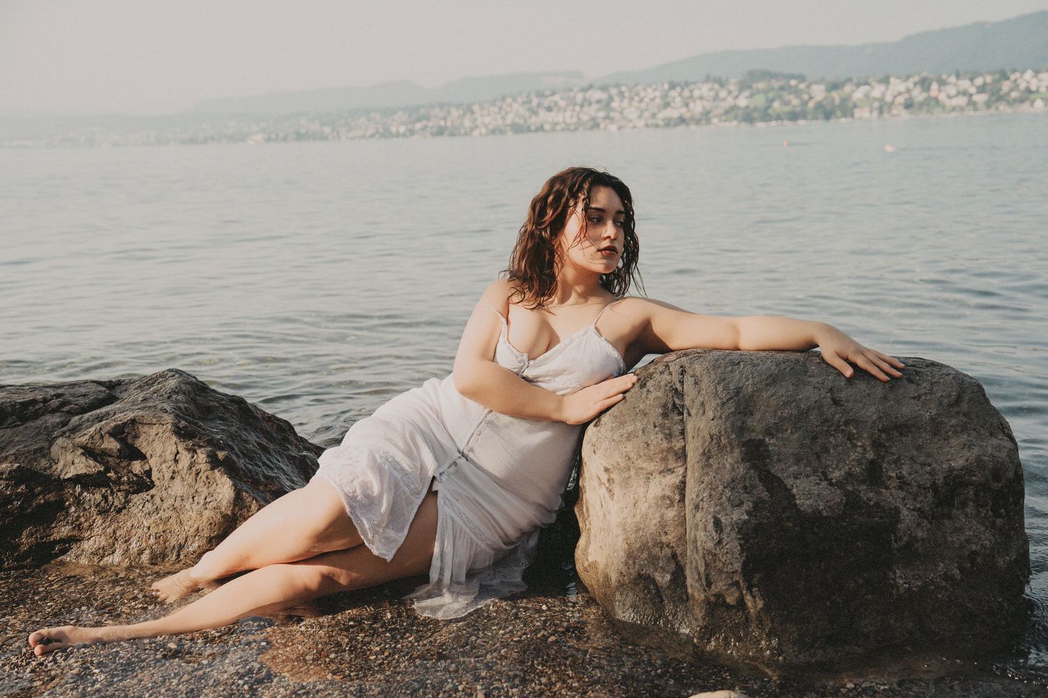 Woman in white dress sitting against a rock by the water.