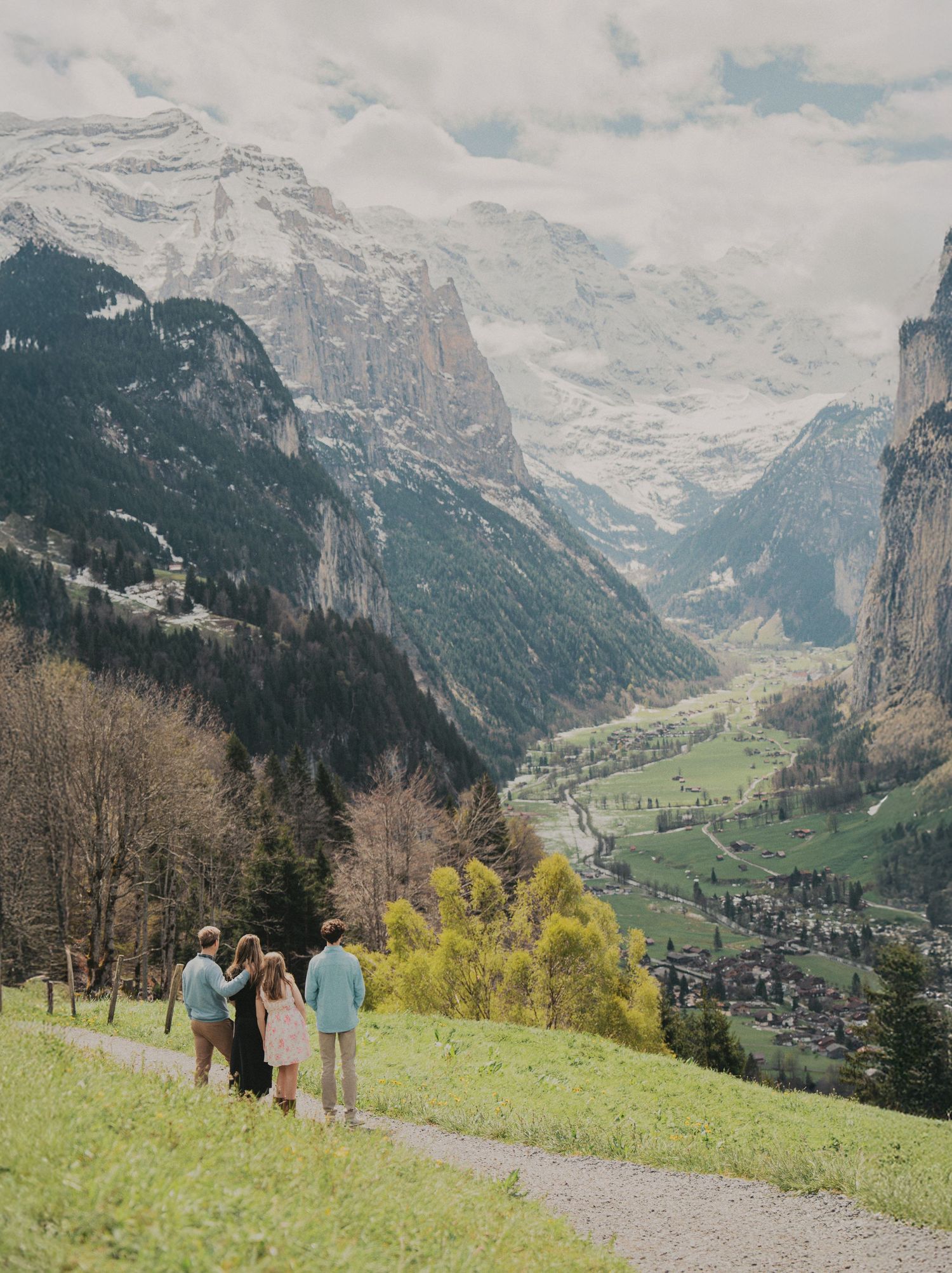 Four family members stand on a hillside path admiring a breathtaking panoramic Swiss mountain valley landscape.