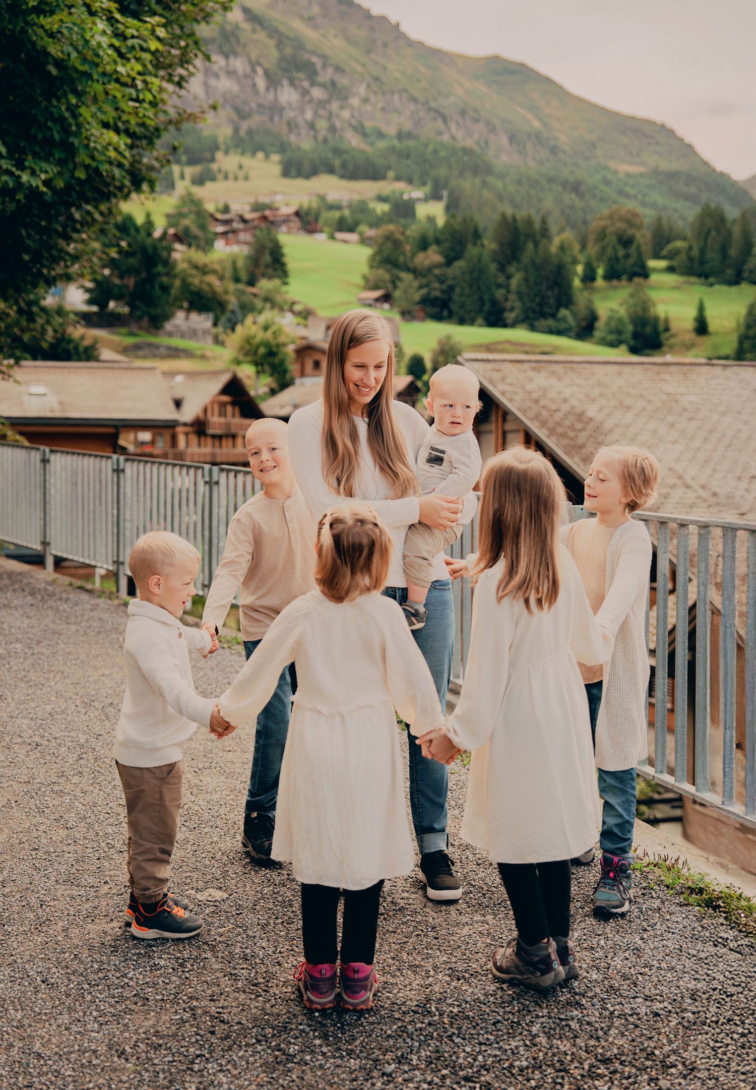 Mother holding toddler surrounded by five children forming a circle on a path in Swiss mountain village.