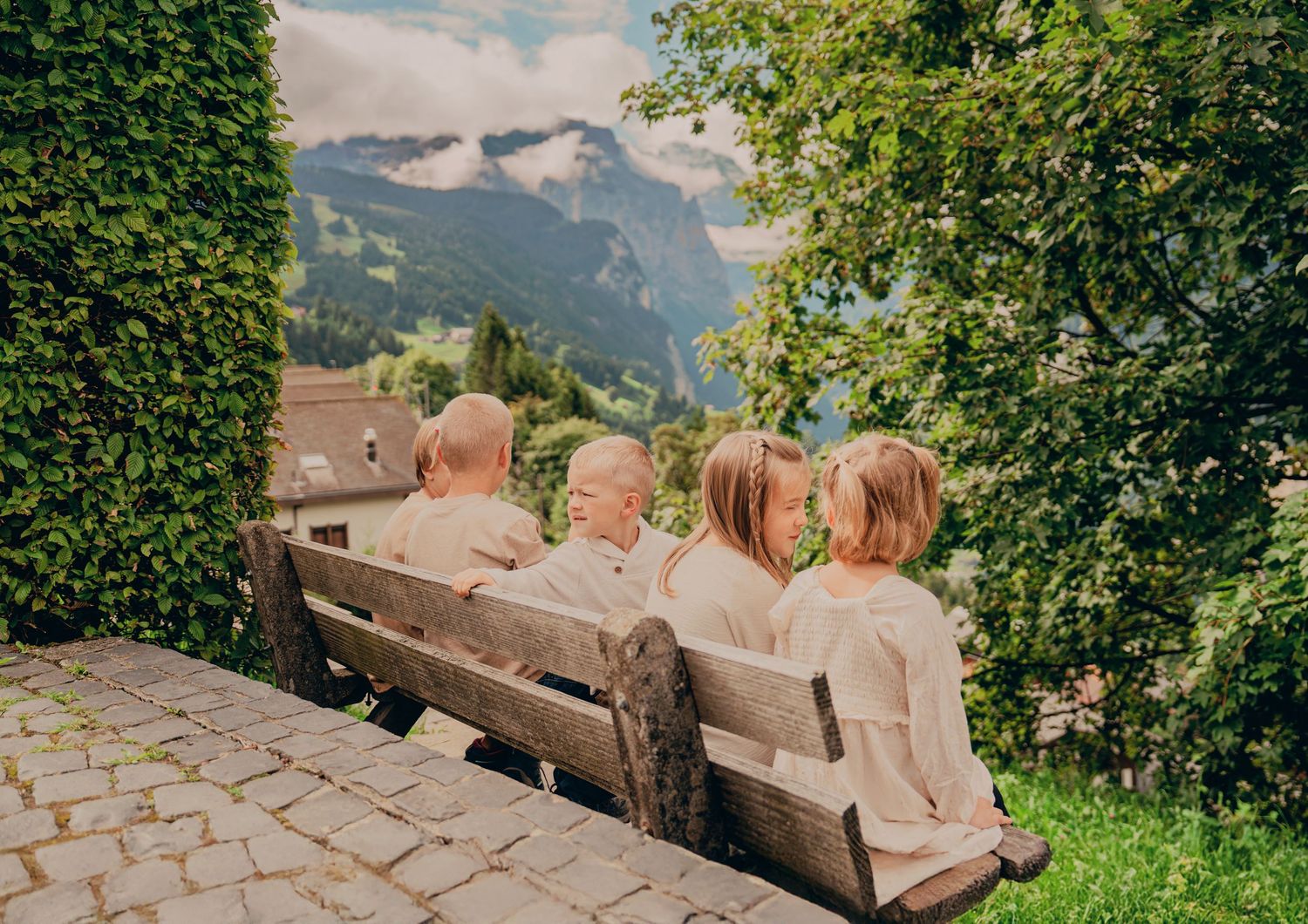 Four young children sitting on a wooden bench viewed from behind, overlooking a stunning Alpine valley landscape.