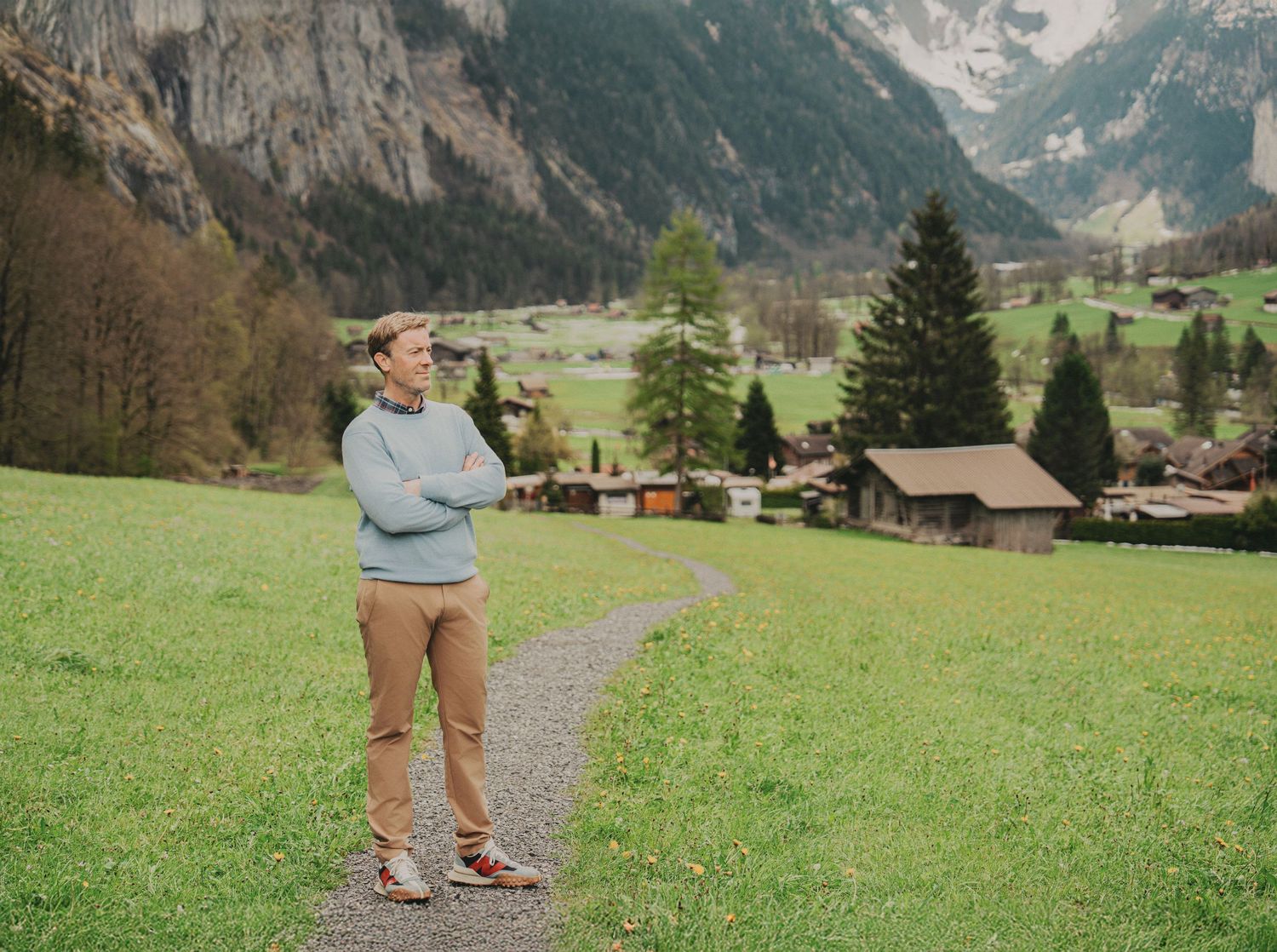 Young man in light blue sweater standing on mountain path overlooking scenic Swiss alpine village.