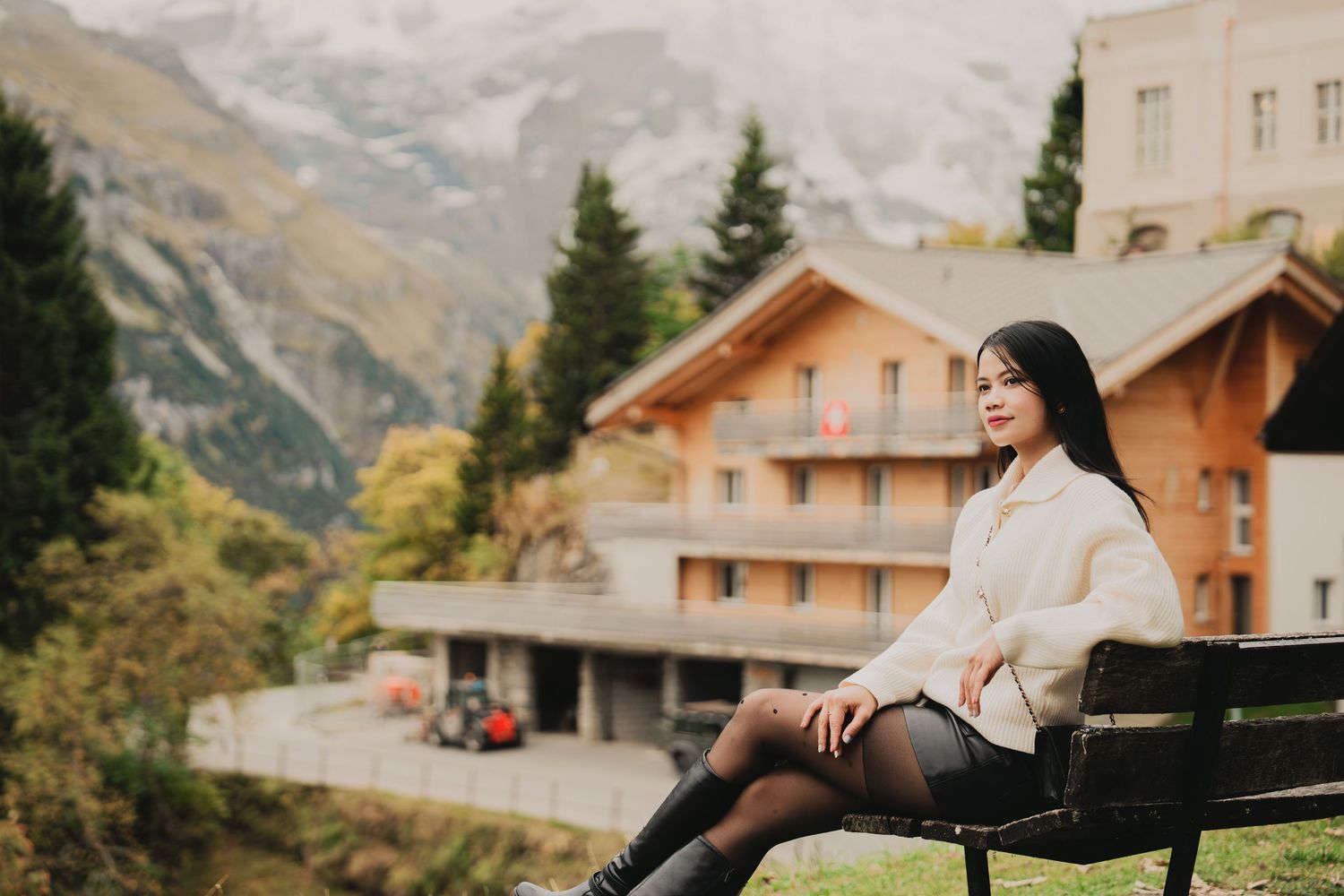 Serene Asian woman in cream sweater and black tights sits on bench near Alpine chalet and mountain scenery.