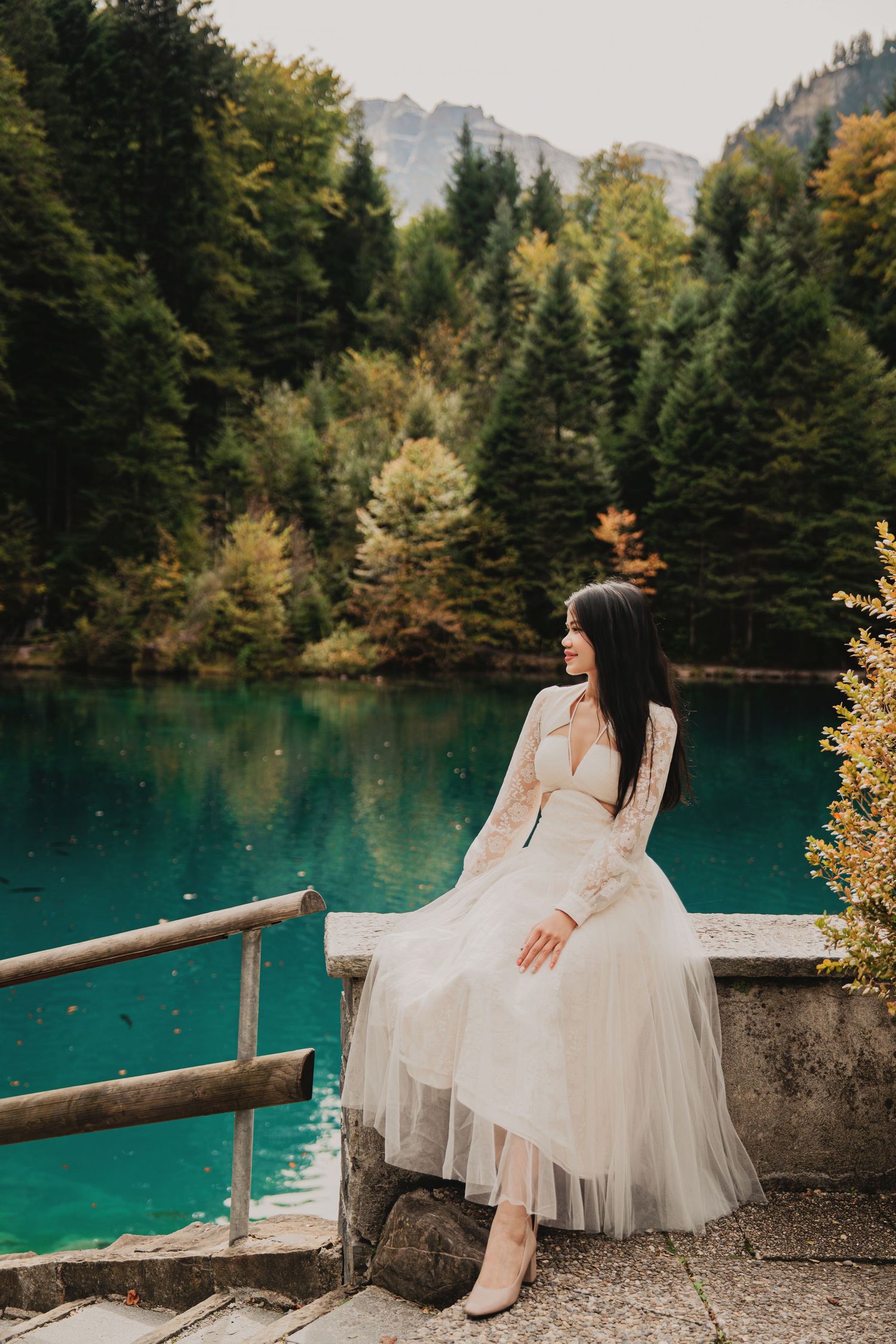 Woman in white tulle dress sits on stone ledge beside tranquil turquoise alpine lake.