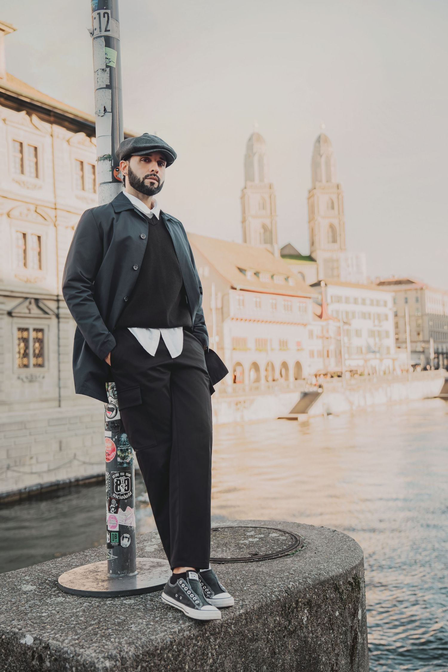 Fashionable bearded man in black coat and flat cap standing by Zurich river with Grossmunster backdrop.