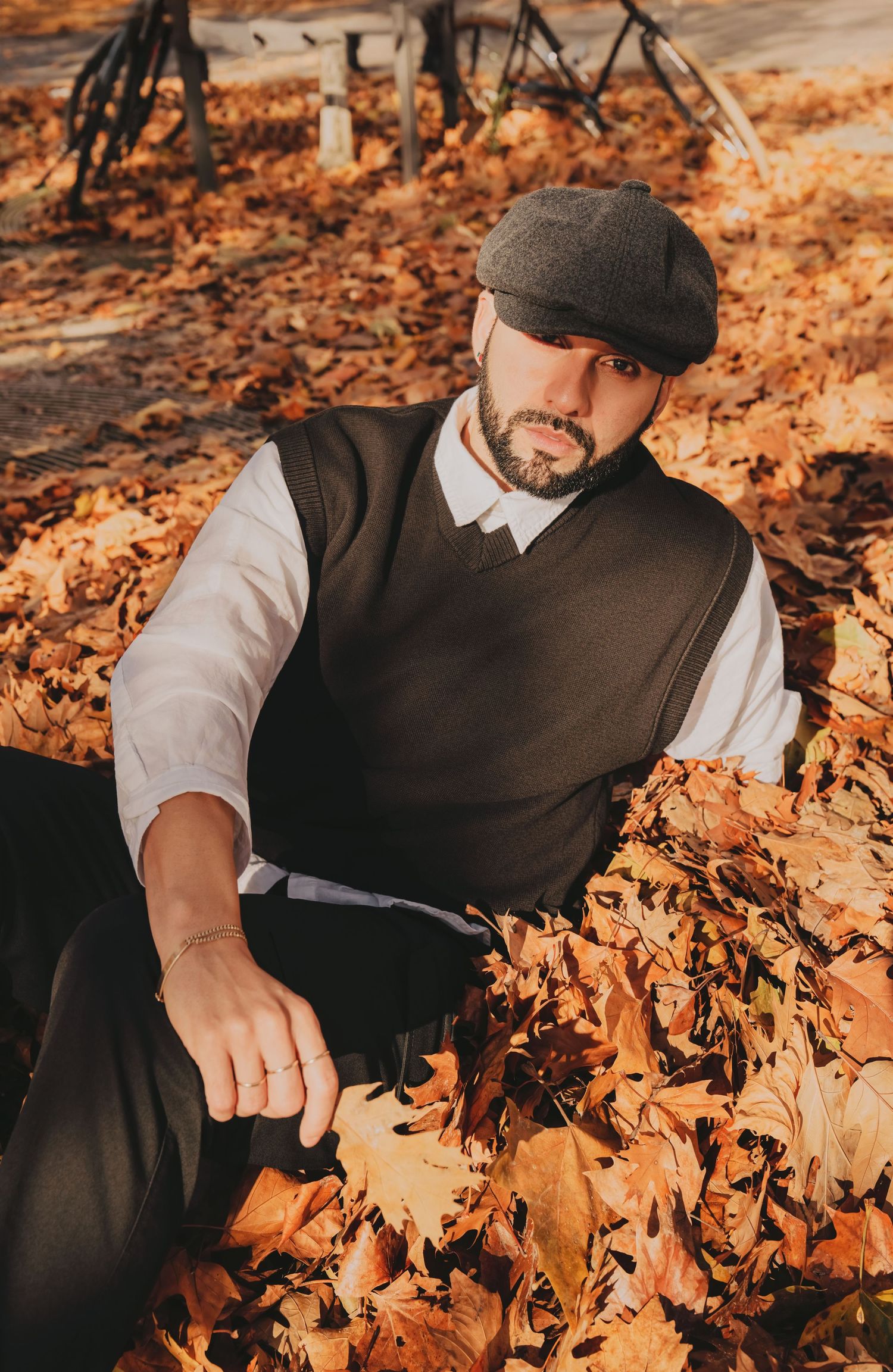Bearded man in dark vest and flat cap sitting relaxed among golden autumn leaves outdoors.