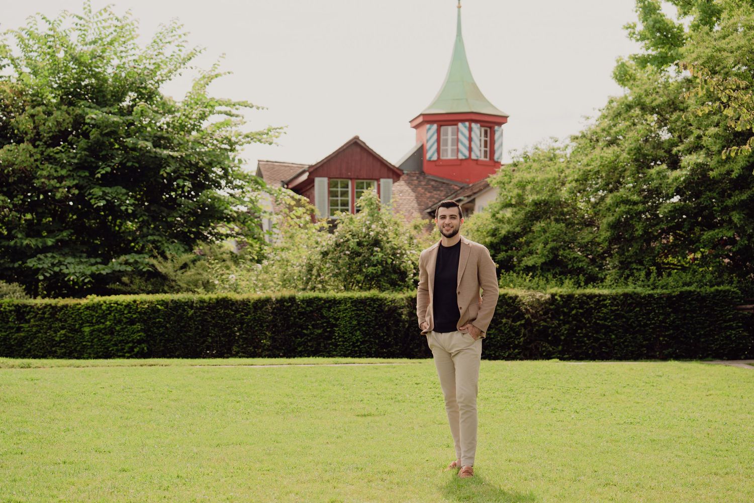 Man in beige blazer standing on green lawn with red-roofed building and lush hedges in background.