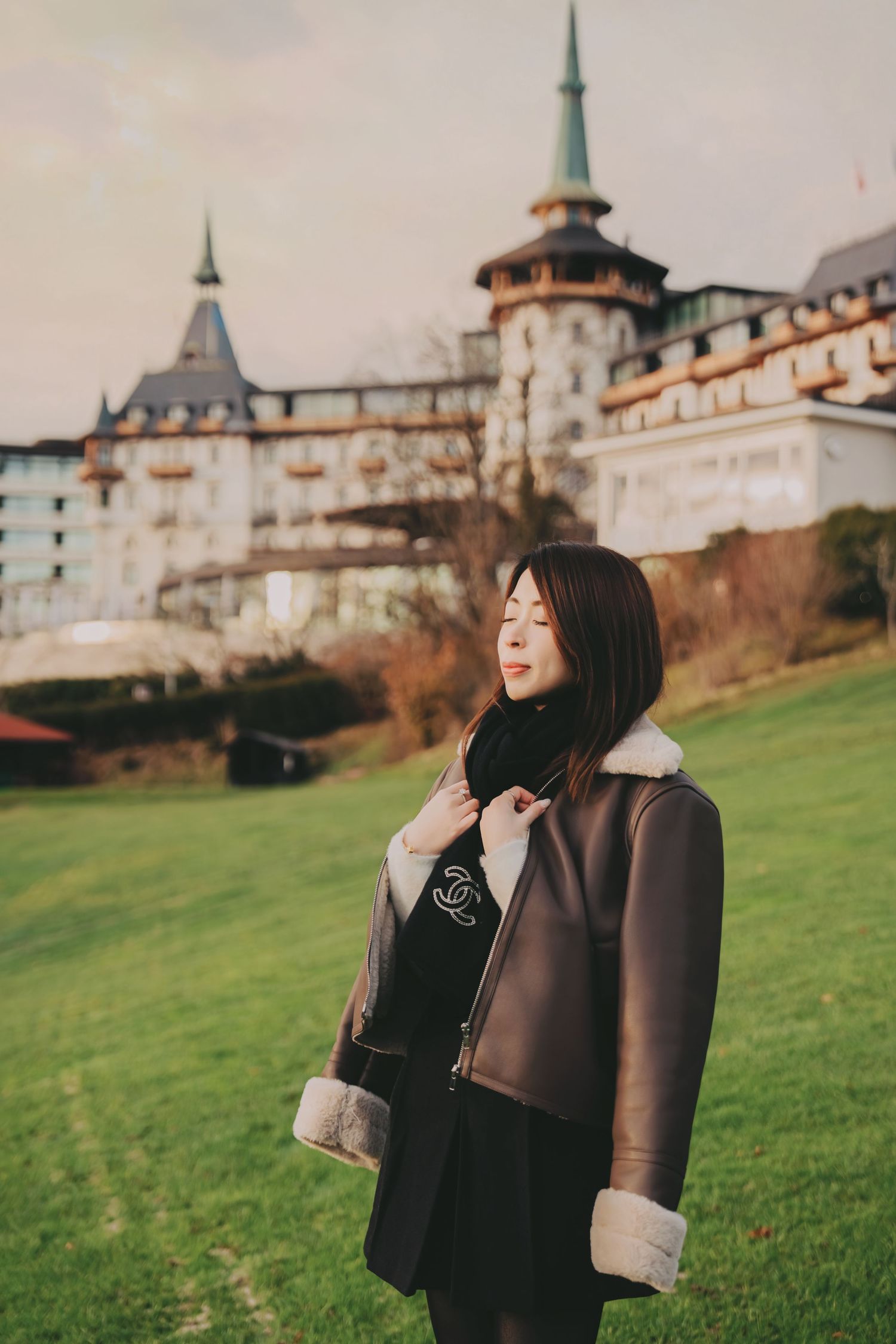 Asian woman in Chanel shearling jacket posing with eyes closed before grand European castle.