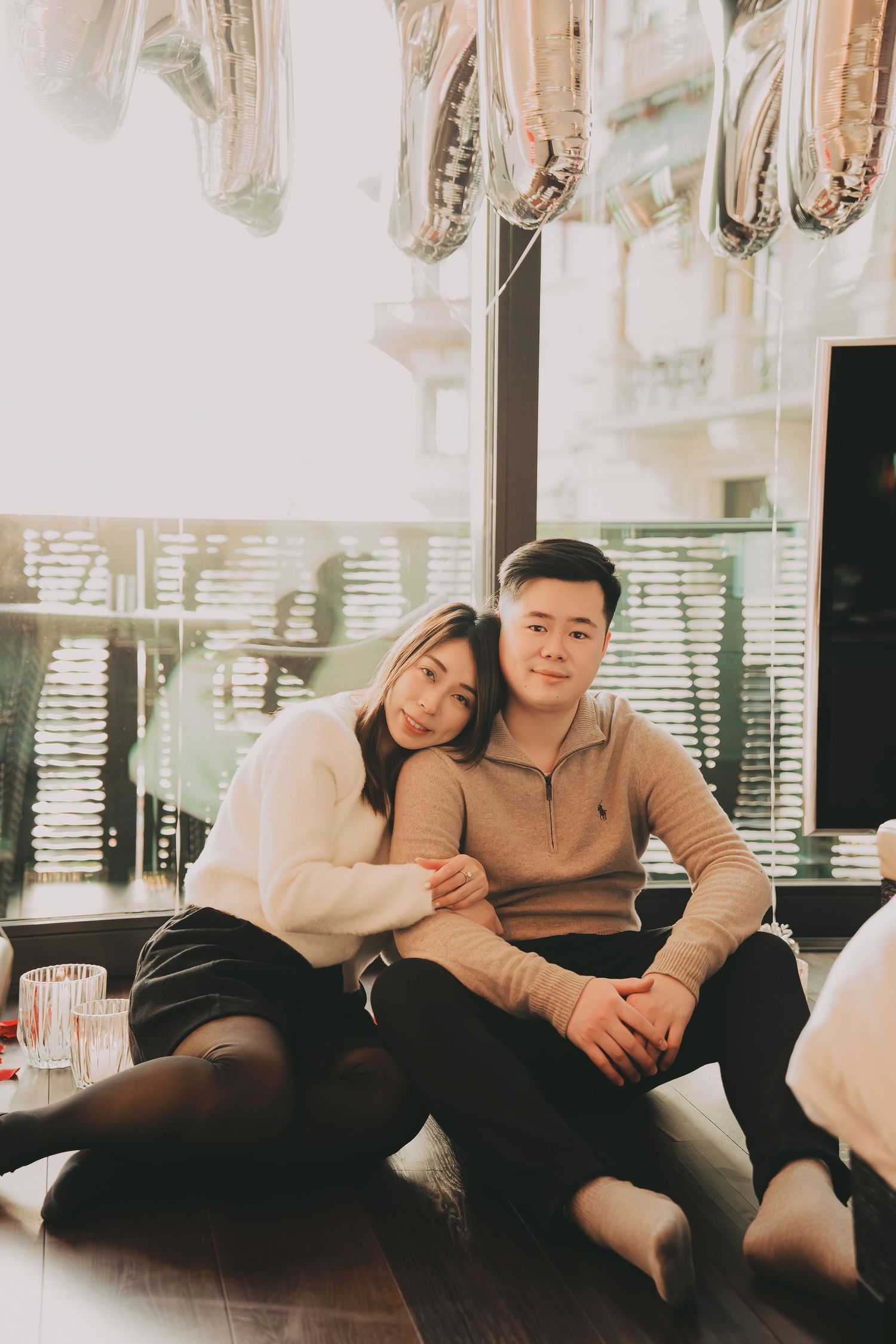 Asian couple sitting on the floor embracing under silver letter balloons in a romantic setting.