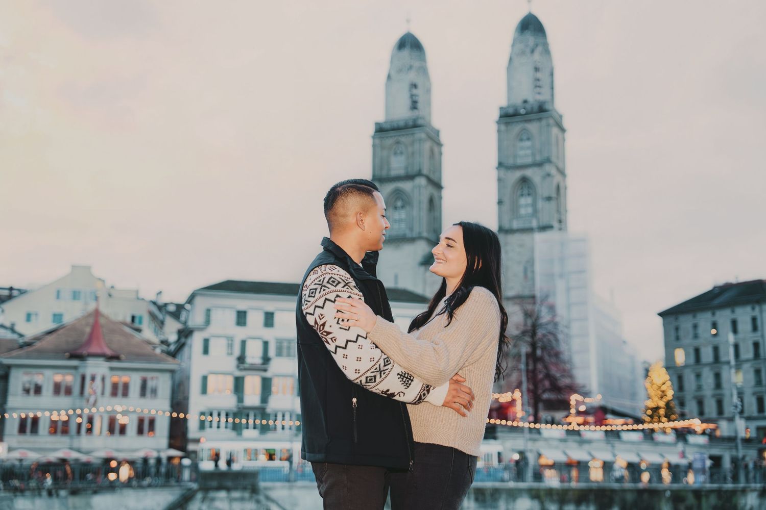 Couple embracing with Grossmünster cathedral and Christmas market lights in Zurich at dusk.