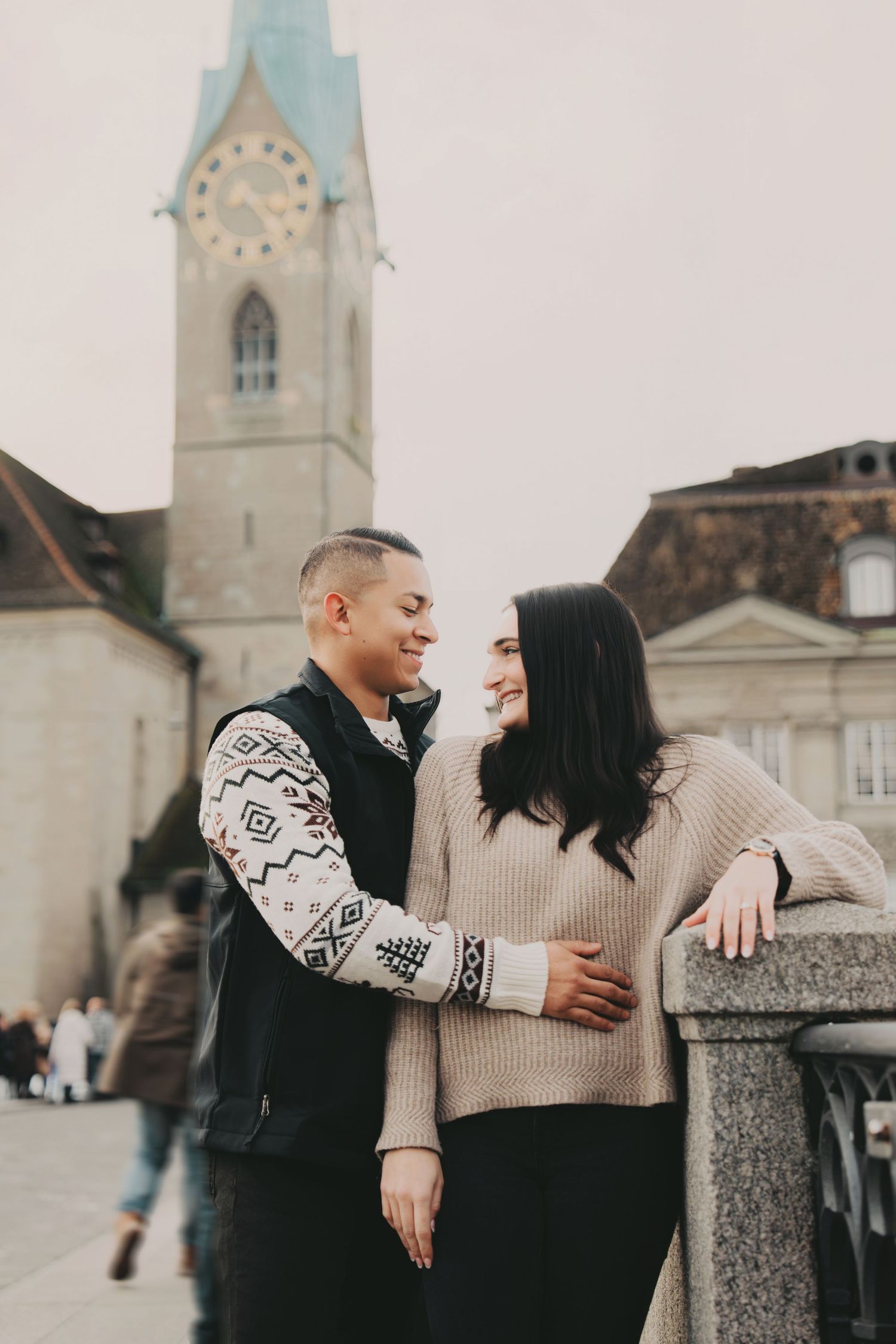 Loving couple smiling at each other on a bridge near a historic clock tower in Zurich, Switzerland.