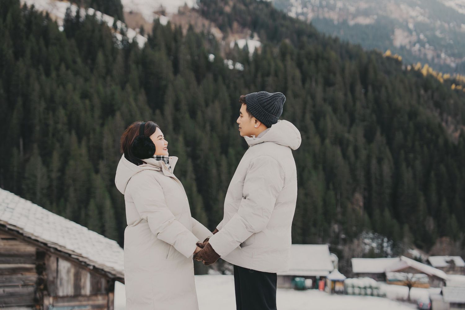 Couple holding hands facing each other in snowy mountain landscape with pine forest background.