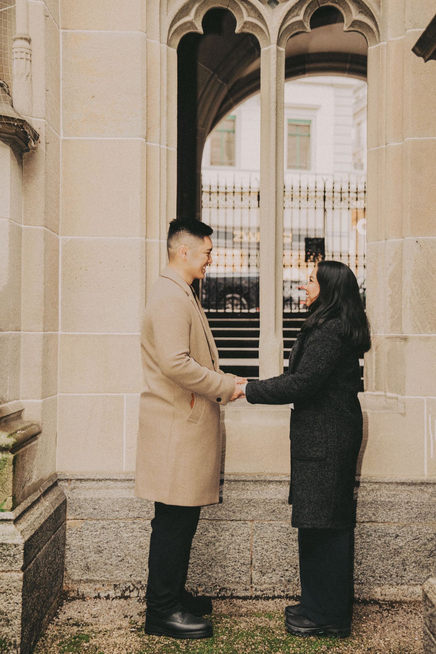 Couple holding hands facing each other outside gothic stone building.