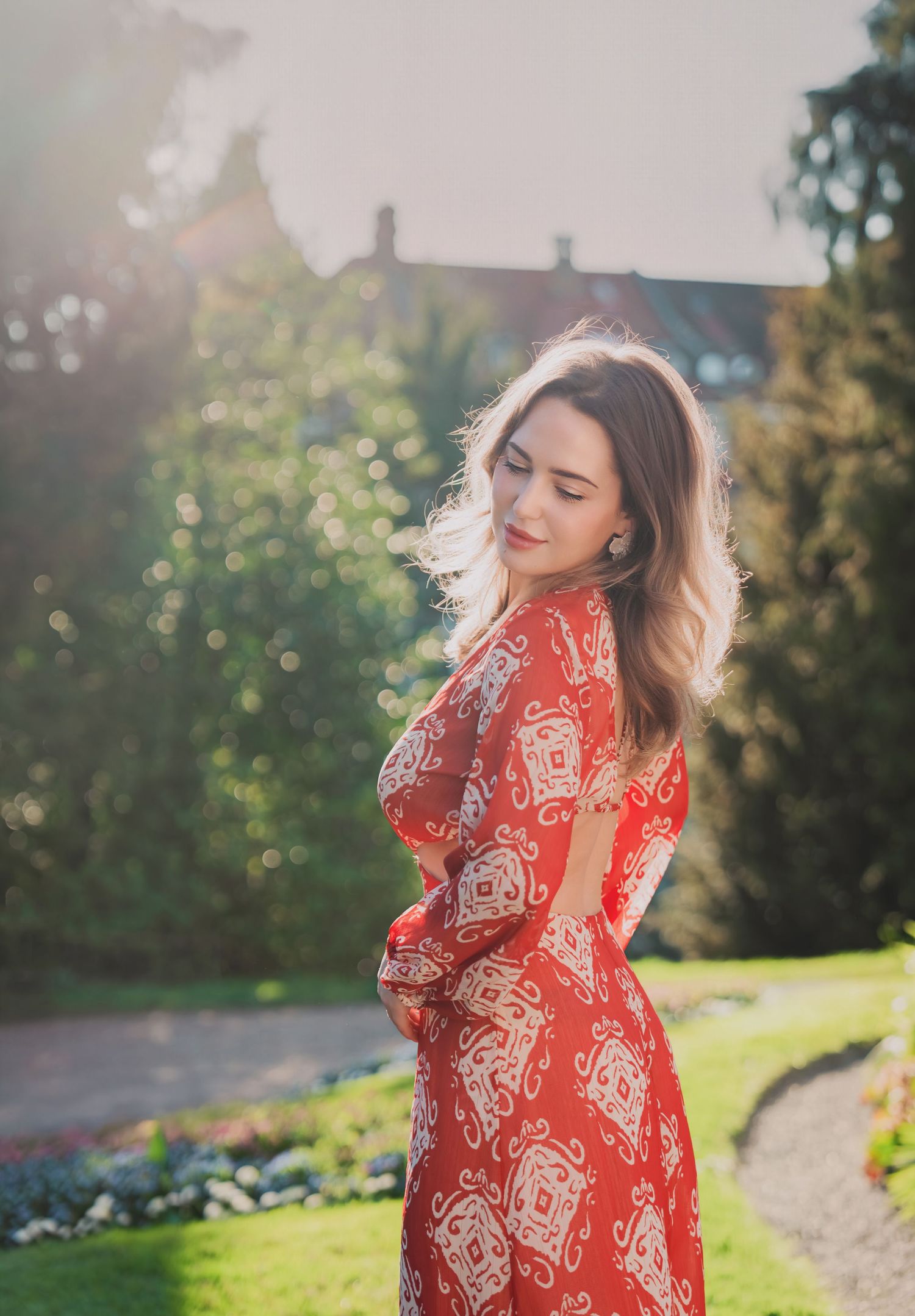 Young woman in red patterned maxi dress poses gracefully in a sunlit garden with lush green trees.