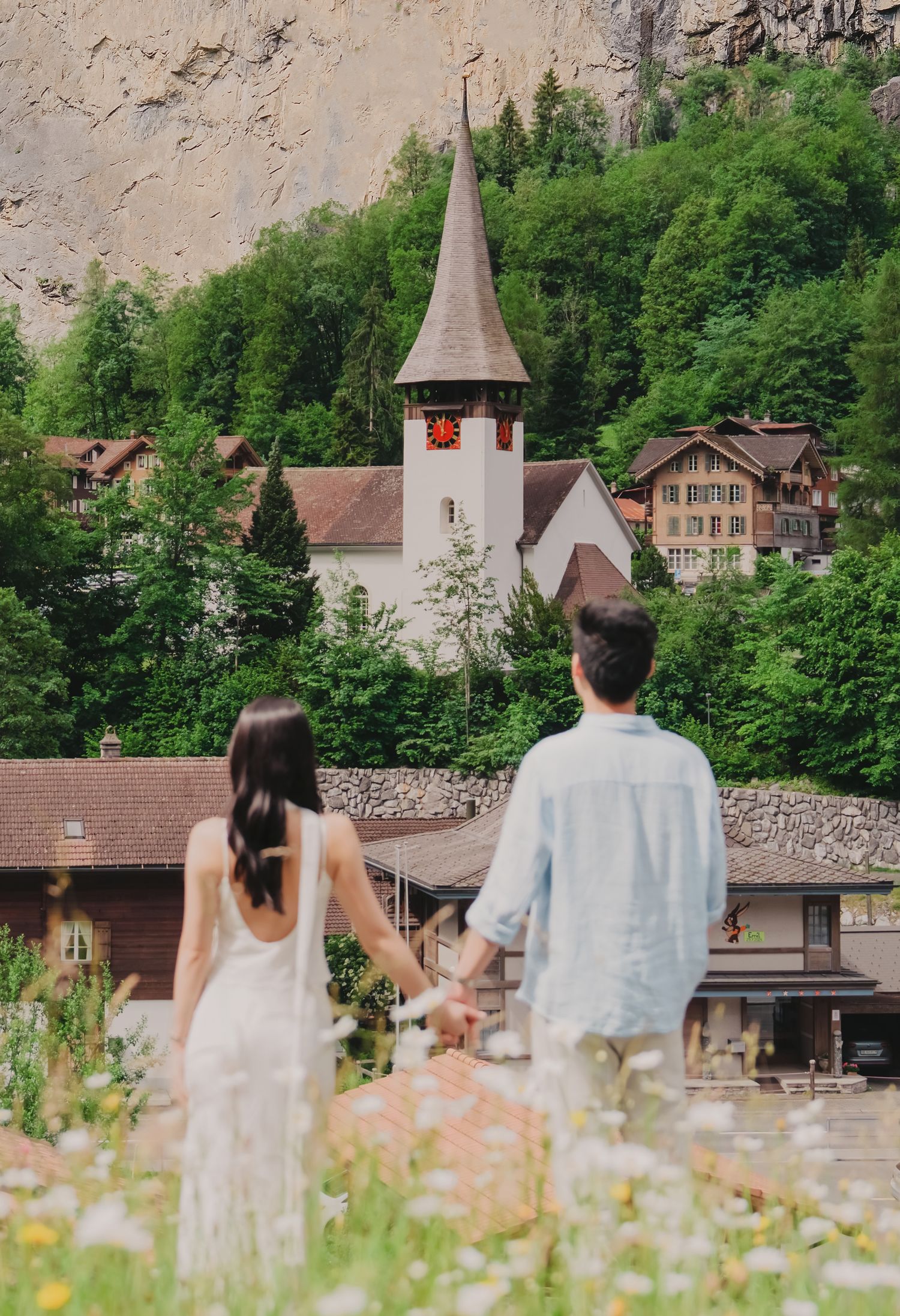 Couple holding hands gazing at a charming Swiss village church surrounded by lush green trees.