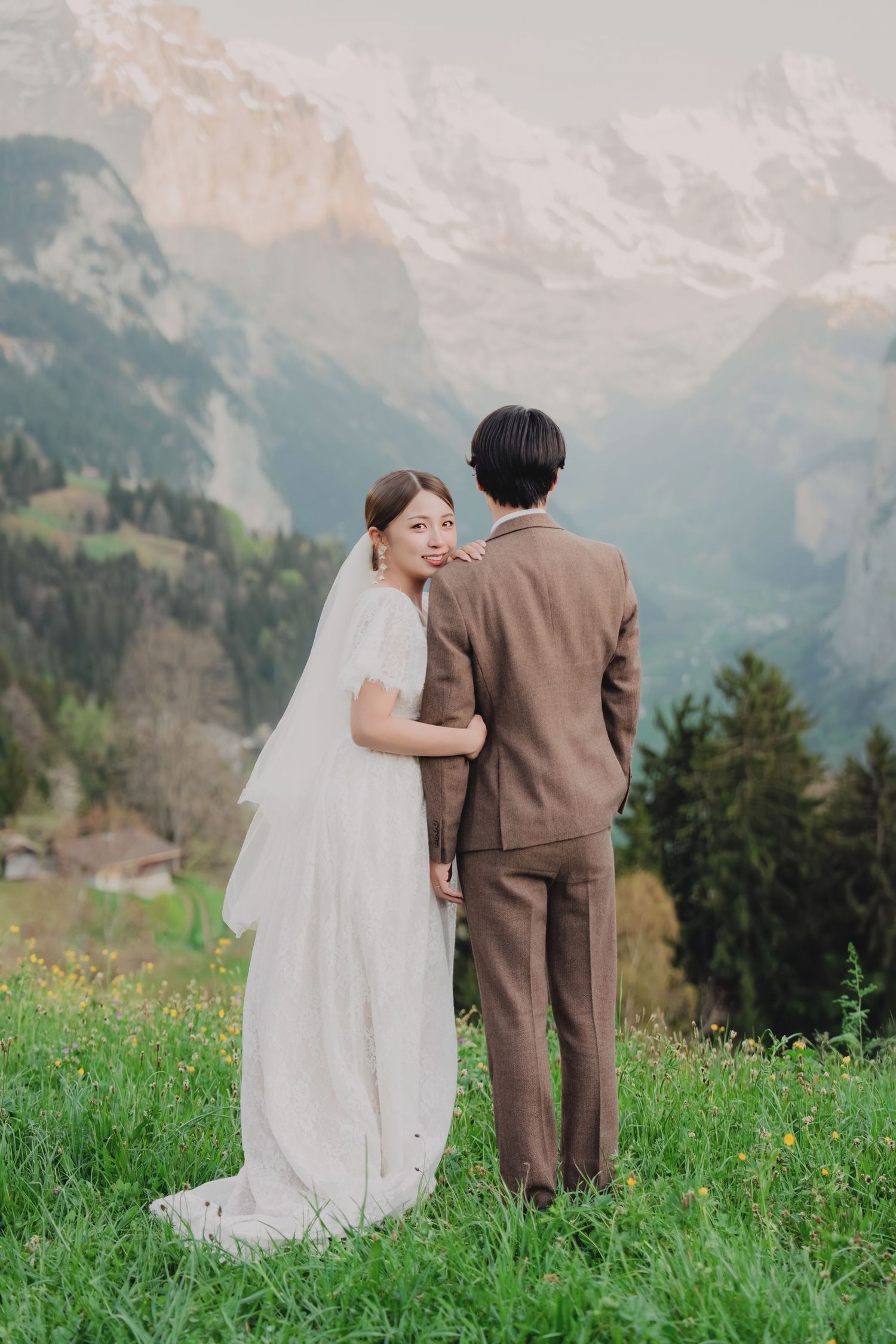 Asian bride in white veil glances back while embracing groom against stunning Swiss alpine mountain backdrop.