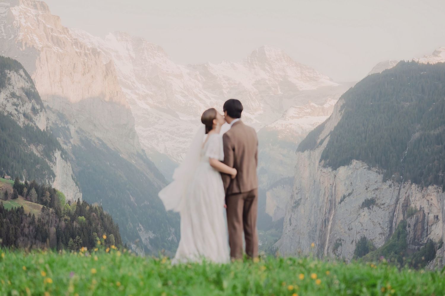 A couple in wedding attire stands embracing on a green meadow with dramatic Alpine mountain views behind them.