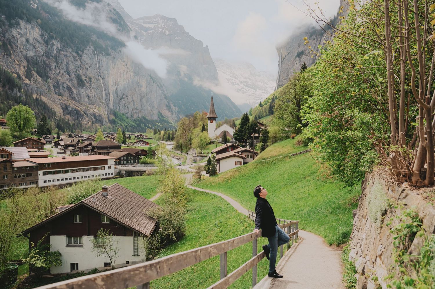 Person standing on scenic overlook path above picturesque Swiss valley village surrounded by mountains.