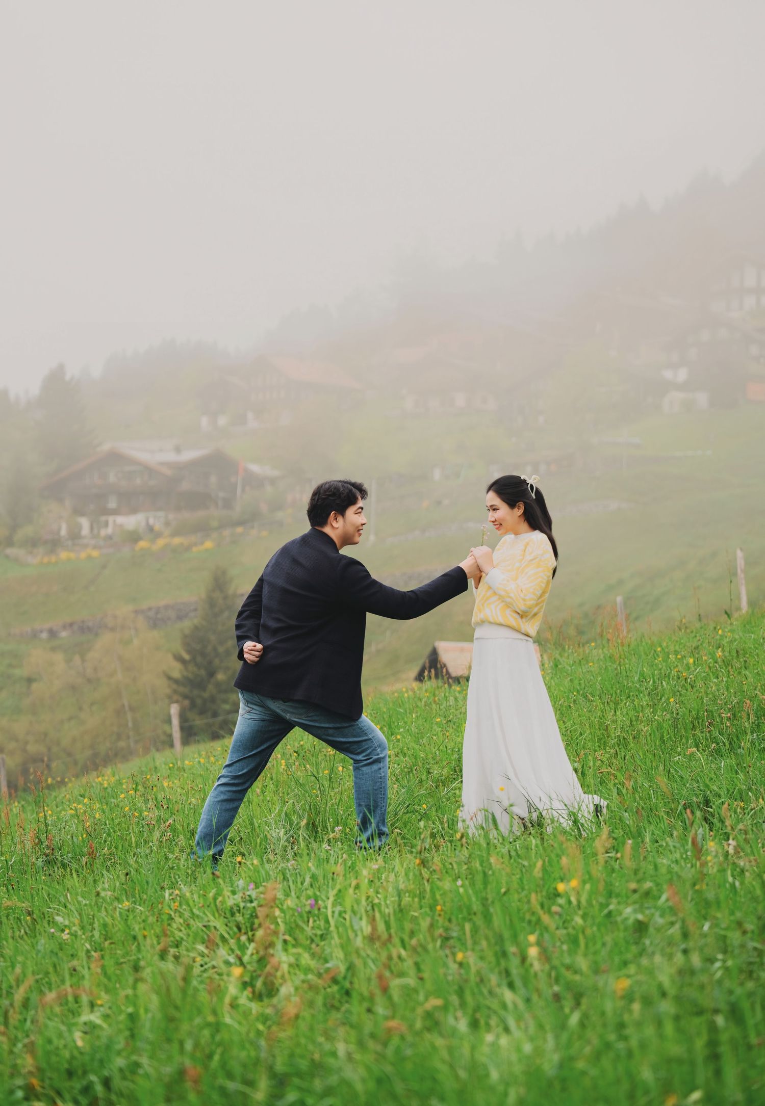 Couple playfully reaching toward each other on a green hillside with misty village backdrop.