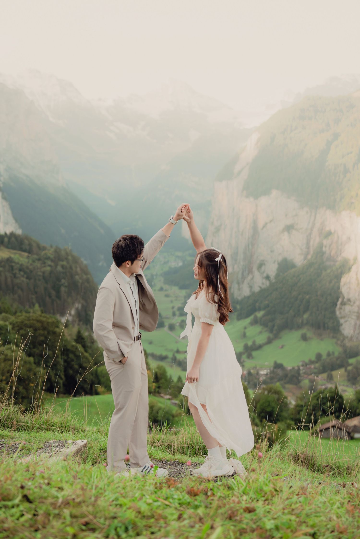 Groom spinning bride in white dress on alpine meadow with breathtaking Swiss mountain valley panorama.