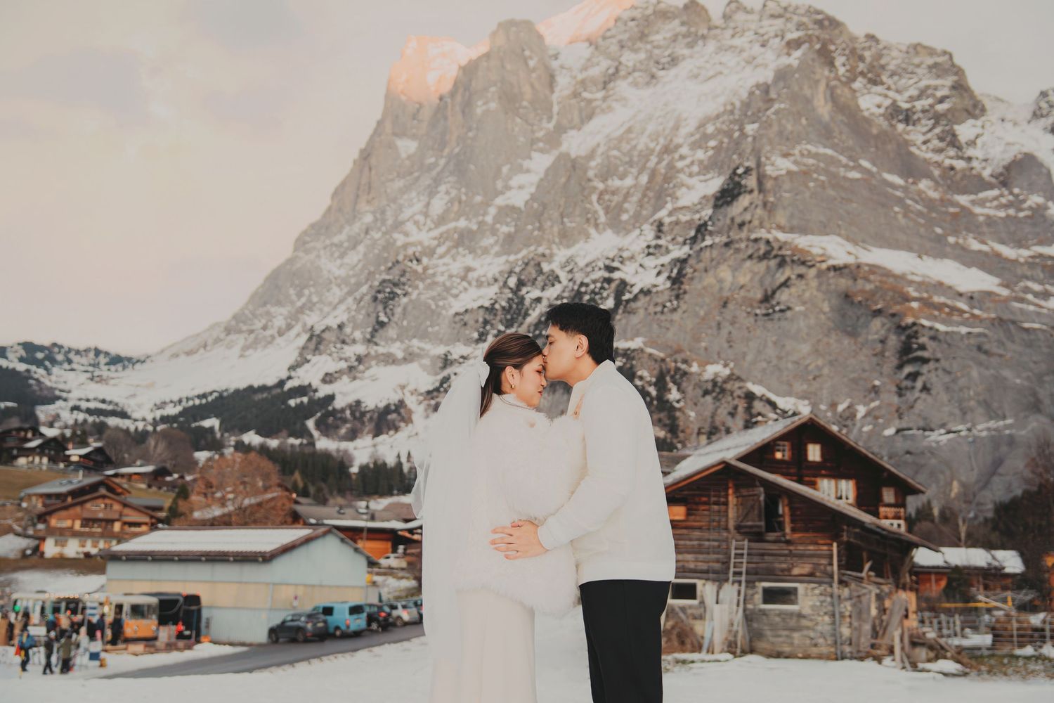 Romantic couple embracing in snowy alpine village with golden sunset illuminating mountain peaks behind them.