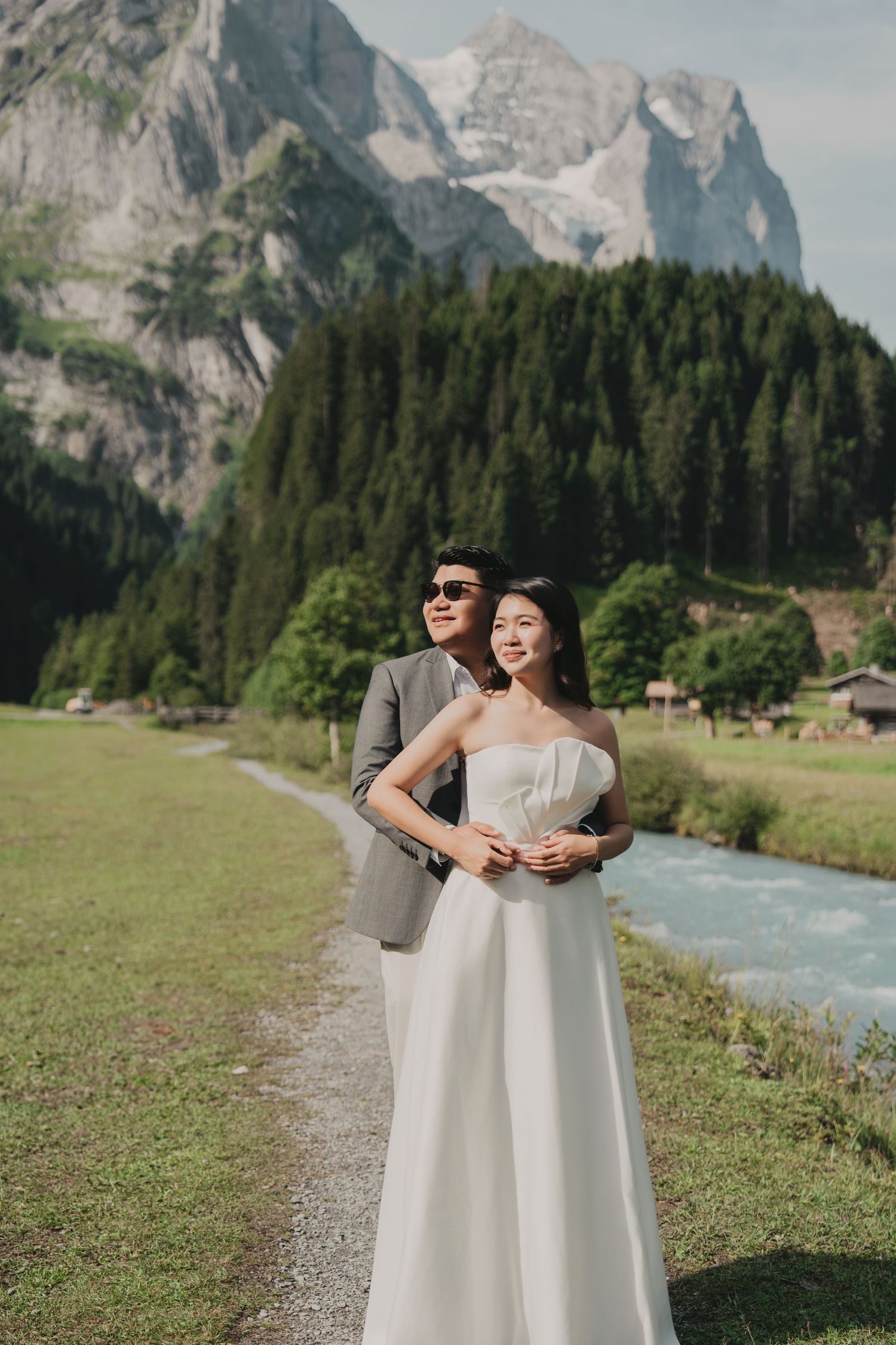 Elegant couple posing on an Alpine path with glacial peaks, pine forest, and stream behind them.