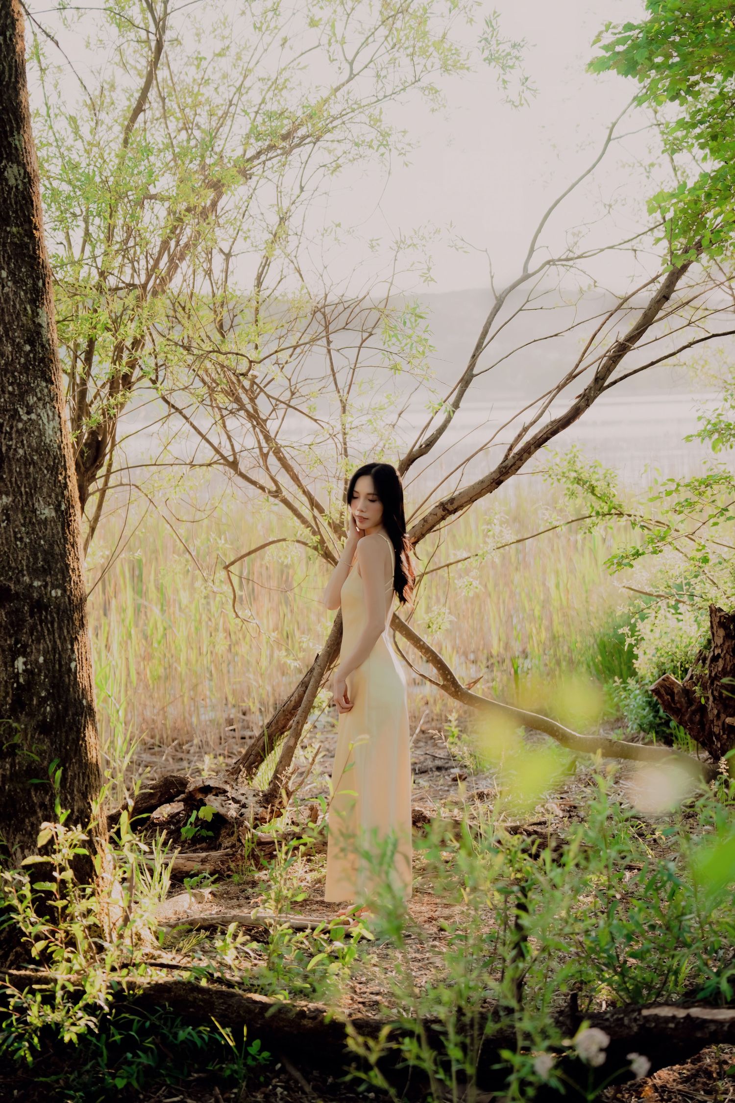 Woman in elegant cream dress standing among trees near water in a serene natural setting.