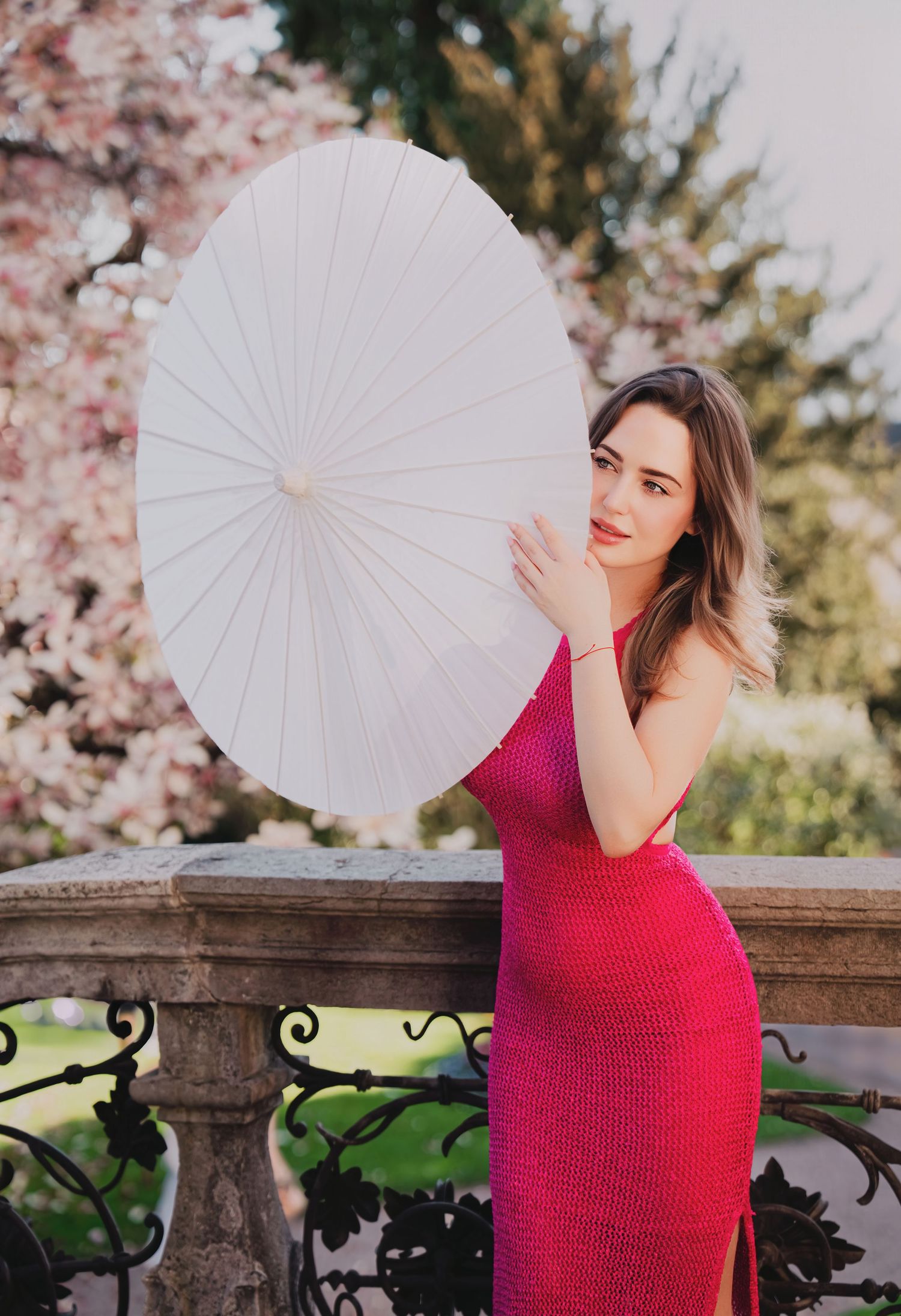 Woman in pink dress holding white parasol near cherry blossoms.