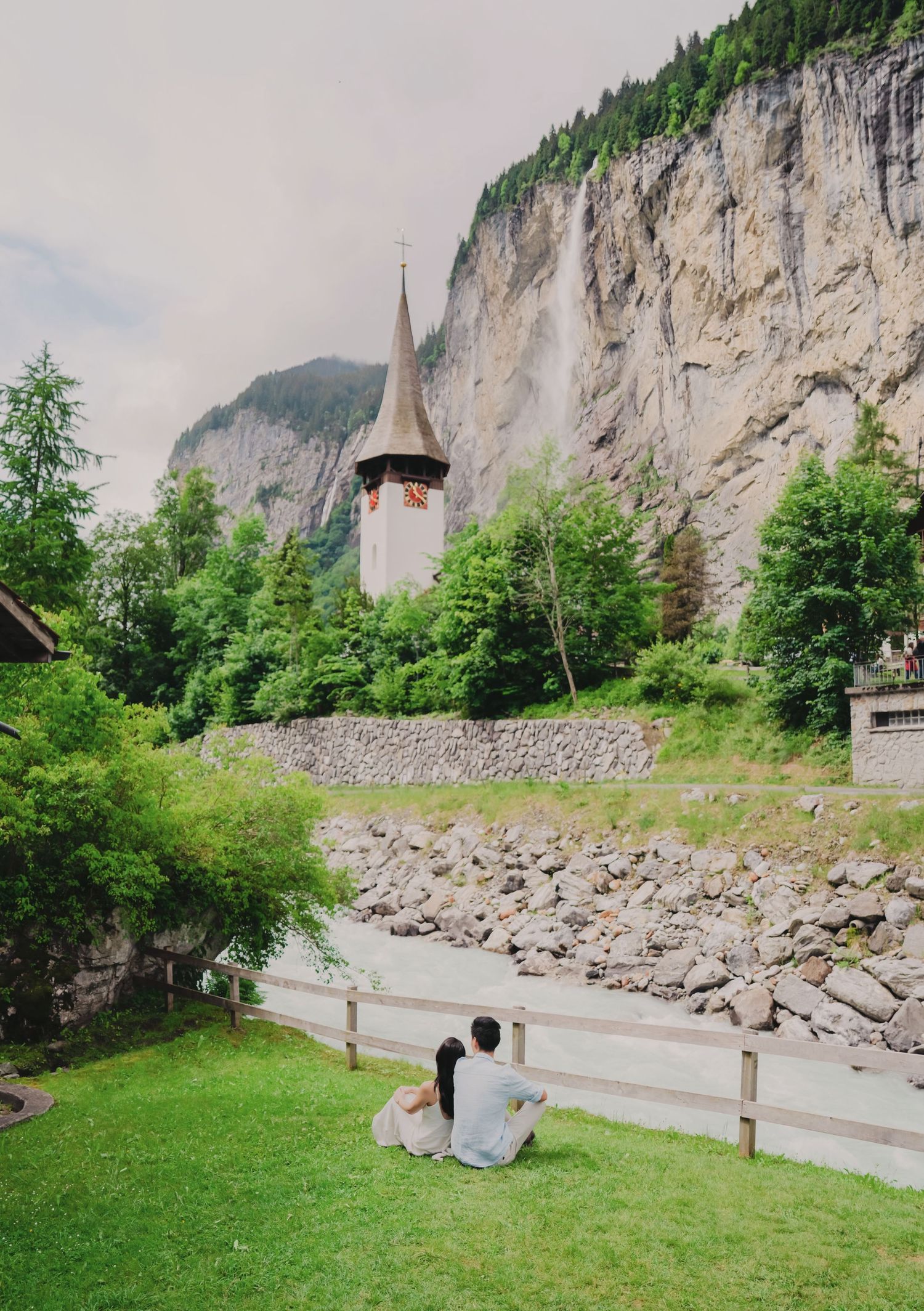Couple sitting together on grass admiring a historic Swiss church tower beside a rocky cliff.