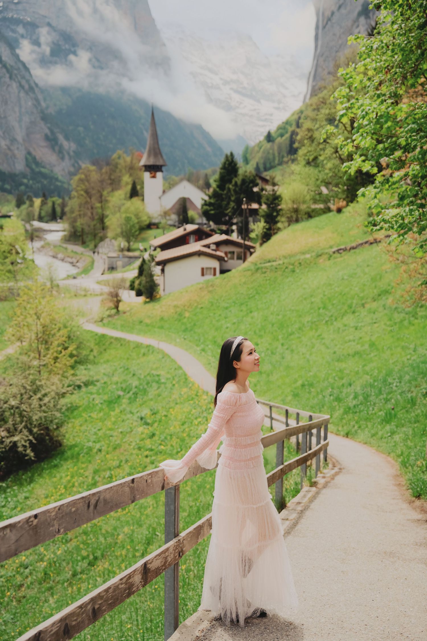 Elegant woman in pink dress standing gracefully by wooden fence on scenic path in lush Swiss mountain village.