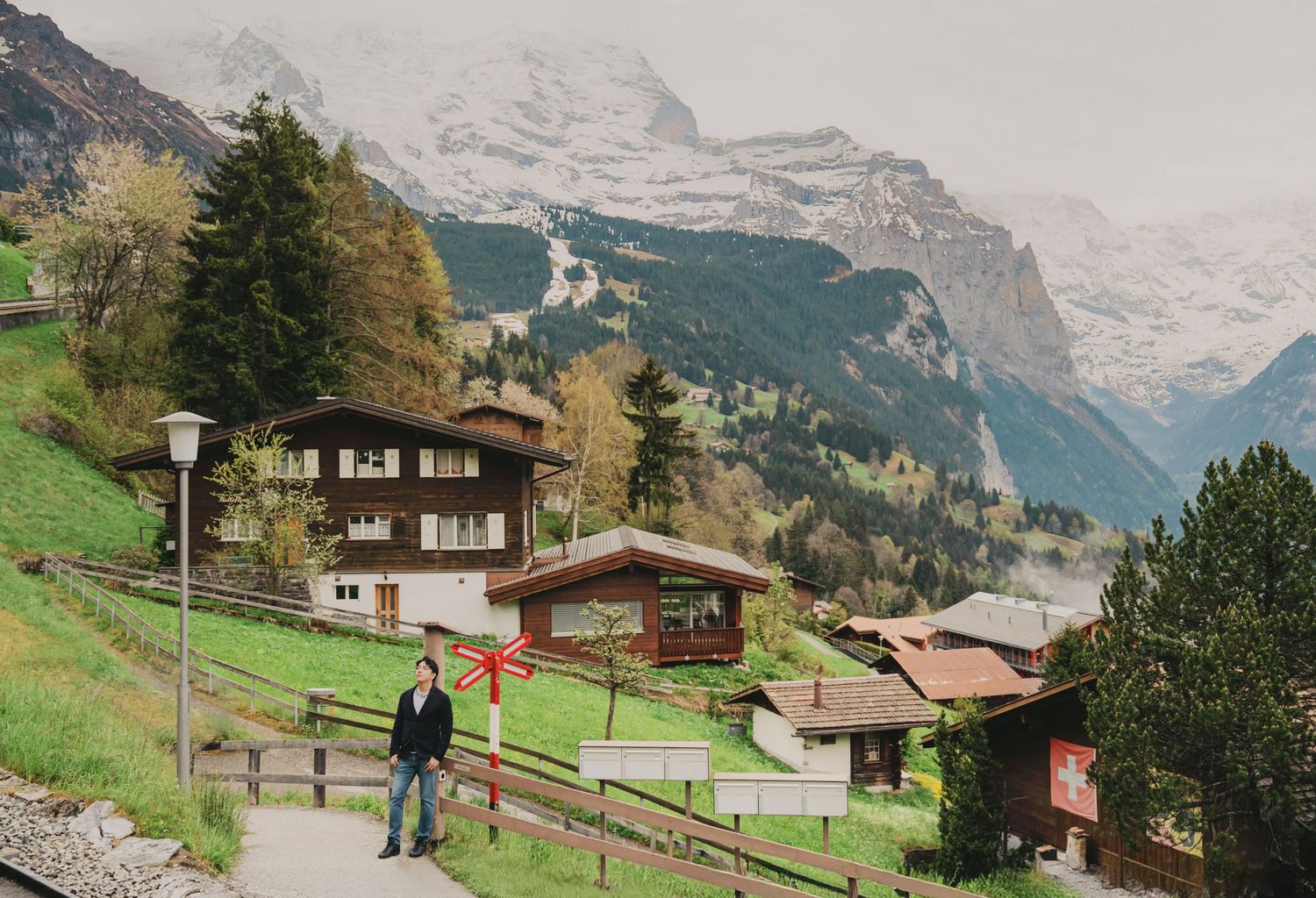 Man standing on a path in a Swiss alpine village with snow-capped mountains and wooden chalets nearby.