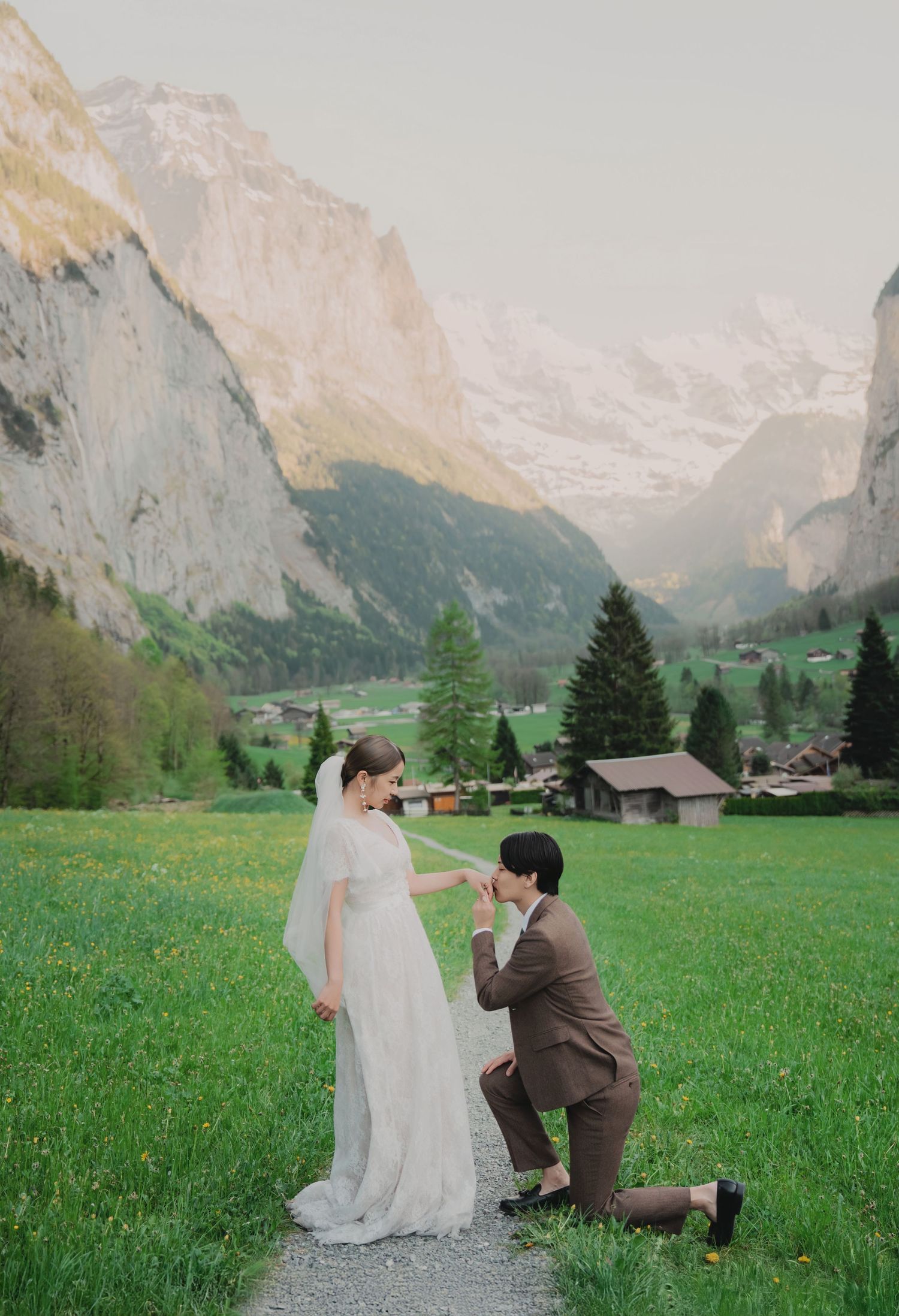 Groom kneeling to kiss bride's hand on a scenic green meadow with majestic Swiss Alps glowing at dusk.
