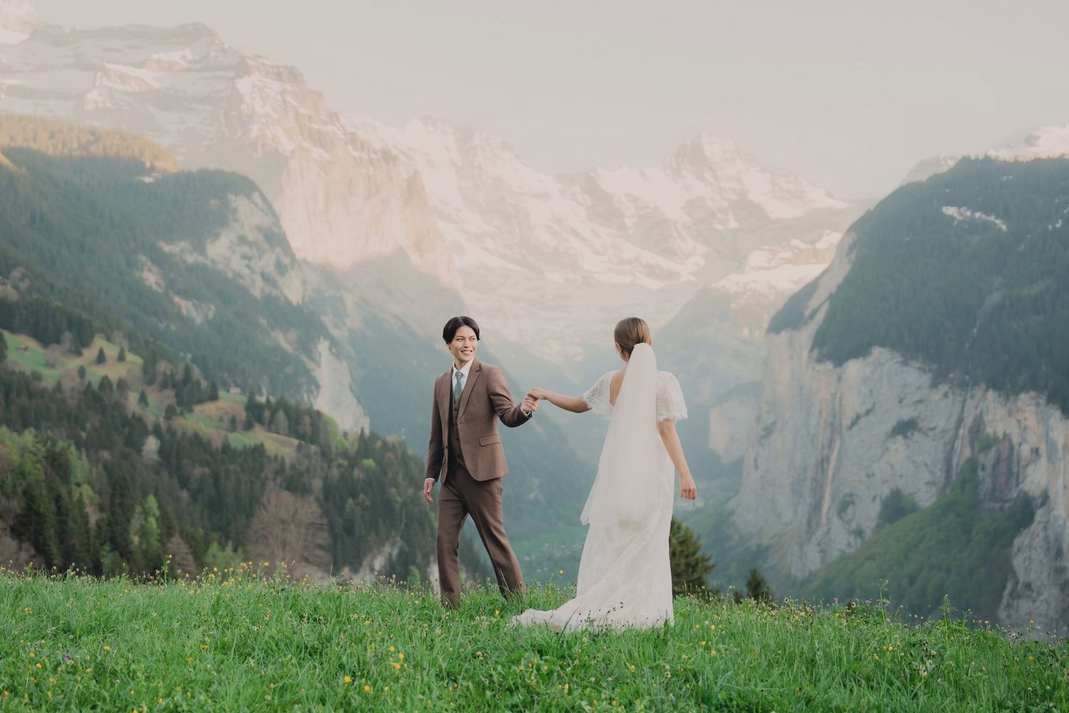 Couple in wedding attire holding hands on a green alpine meadow with dramatic Swiss mountain valley backdrop.
