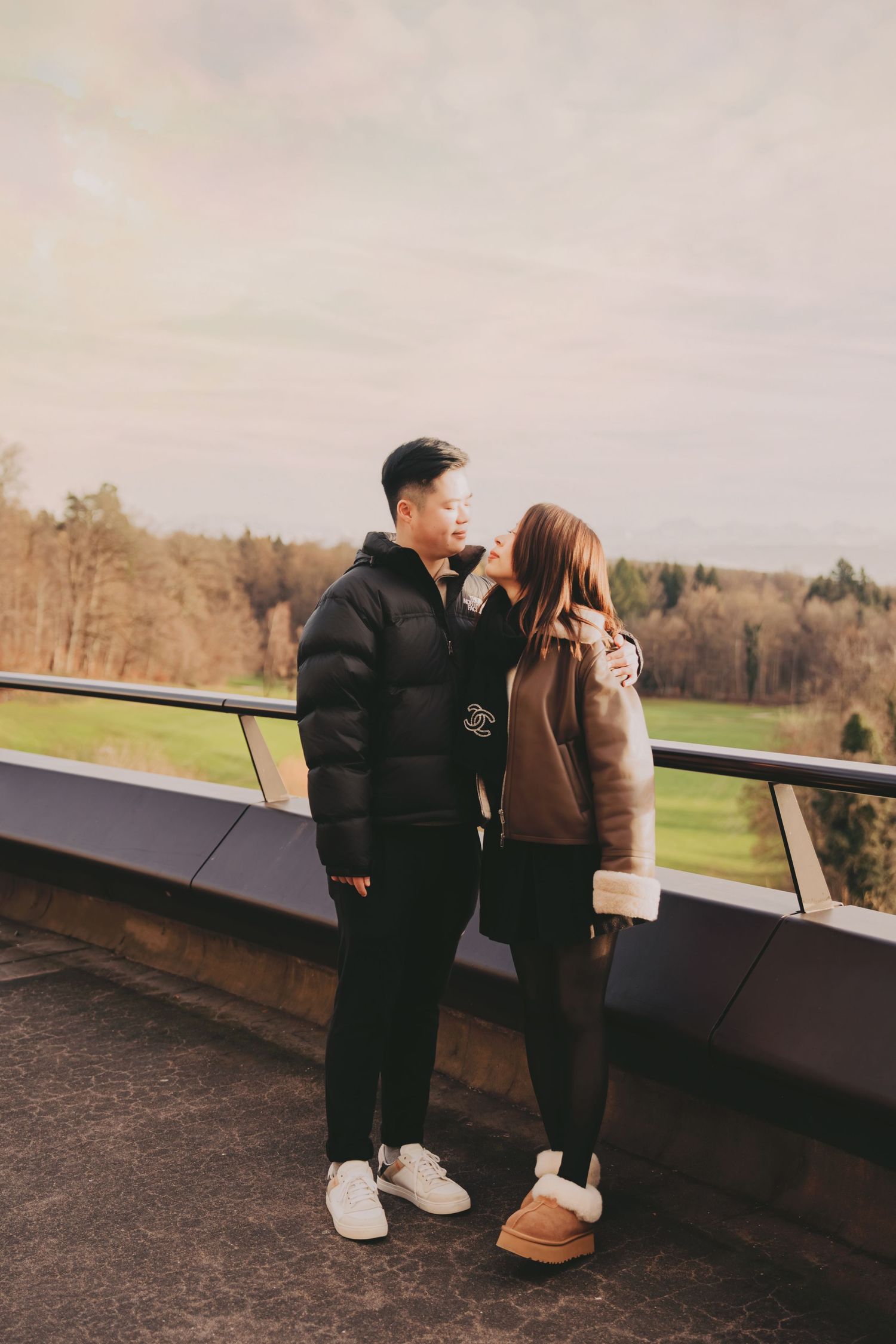 Asian couple standing close together on a balcony overlooking a green countryside landscape.