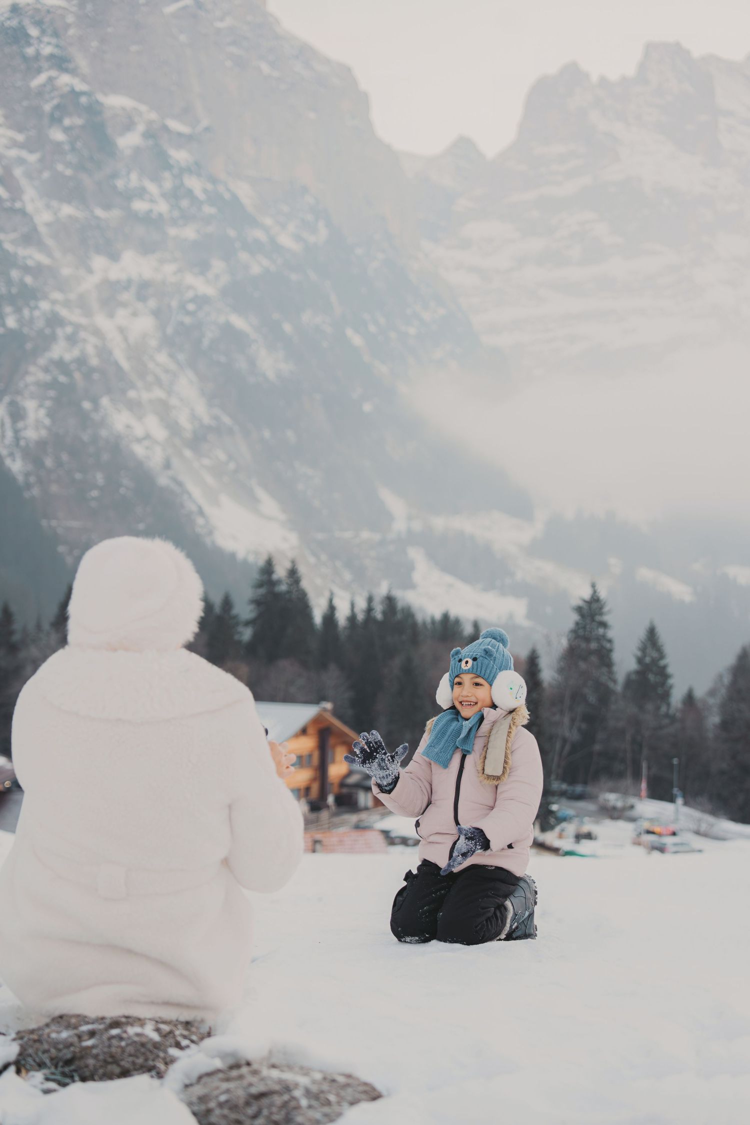 Young girl kneeling in snow interacting with snowman against stunning alpine mountain scenery in winter.