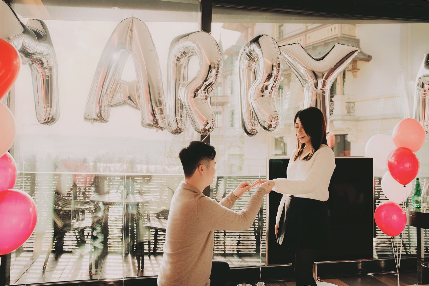 Man proposing to woman with MARRY balloon letters and colorful balloons backdrop.