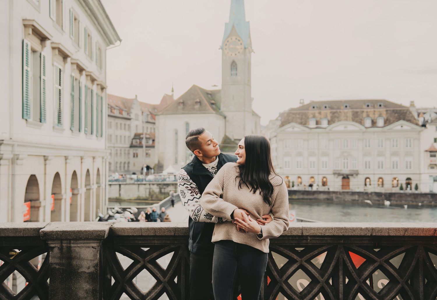 Couple embracing on a bridge overlooking the Limmat River and Fraumunster Church in Zurich.