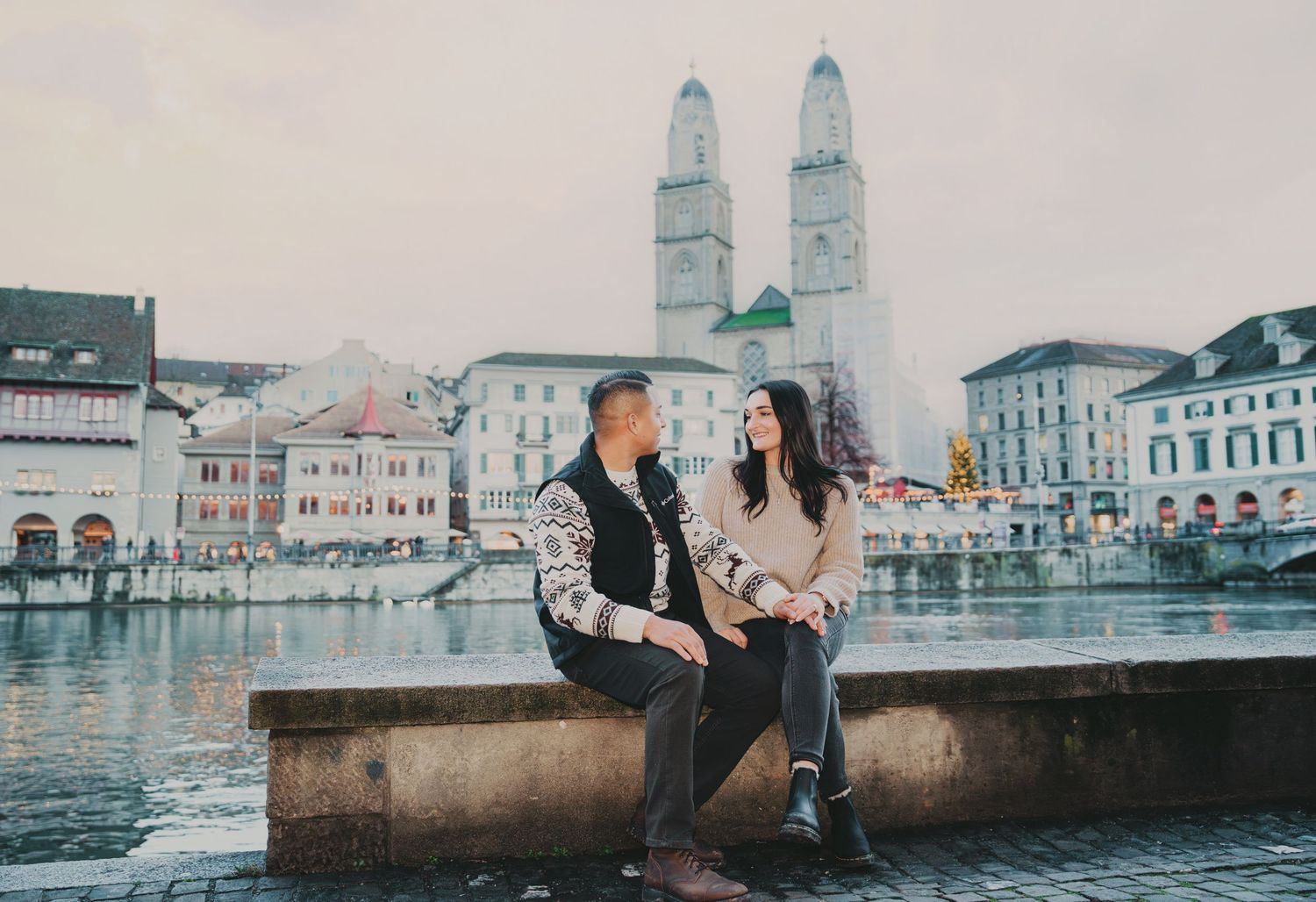 Couple sitting together by the Limmat River with Grossmunster cathedral in the background in Zurich.