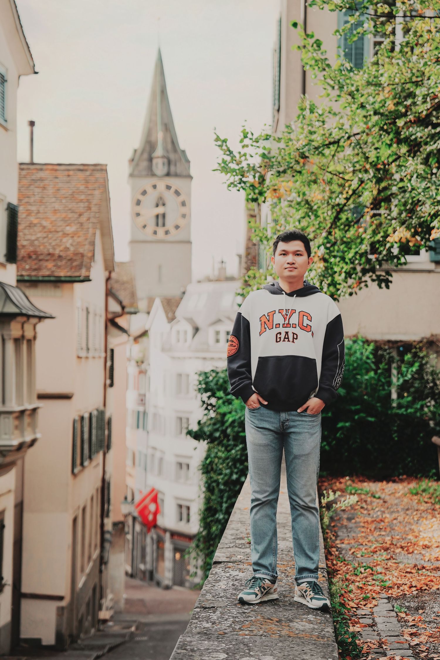 Young man in NYC GAP sweatshirt stands on stone wall in Zurich old town with church tower backdrop.